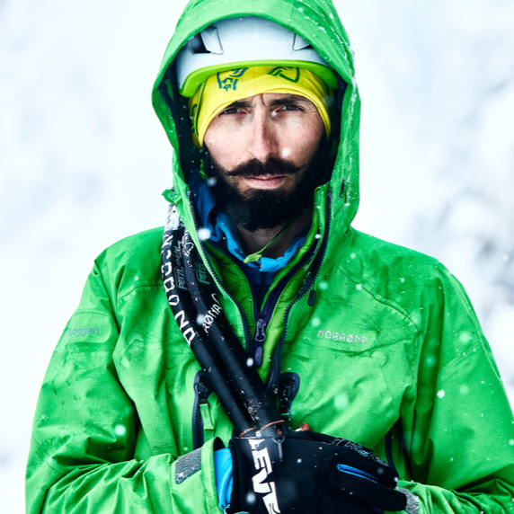 Portrait of a bearded man in snowy conditions wearing a Norrøna green hooded jacket, helmet, and carrying ski poles.
