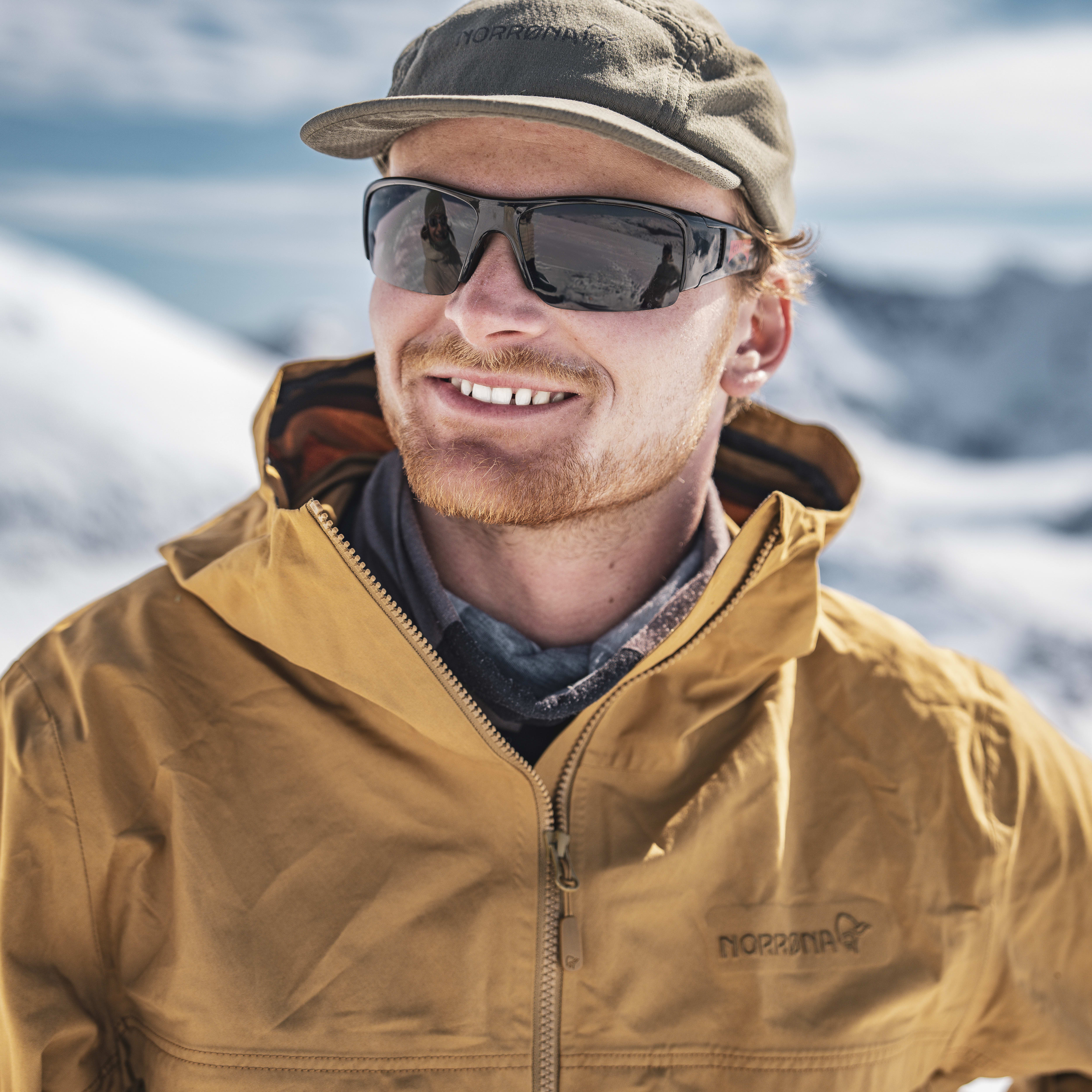 A man wearing a Norrøna jacket and sunglasses smiles while standing in a snowy mountain landscape. 
