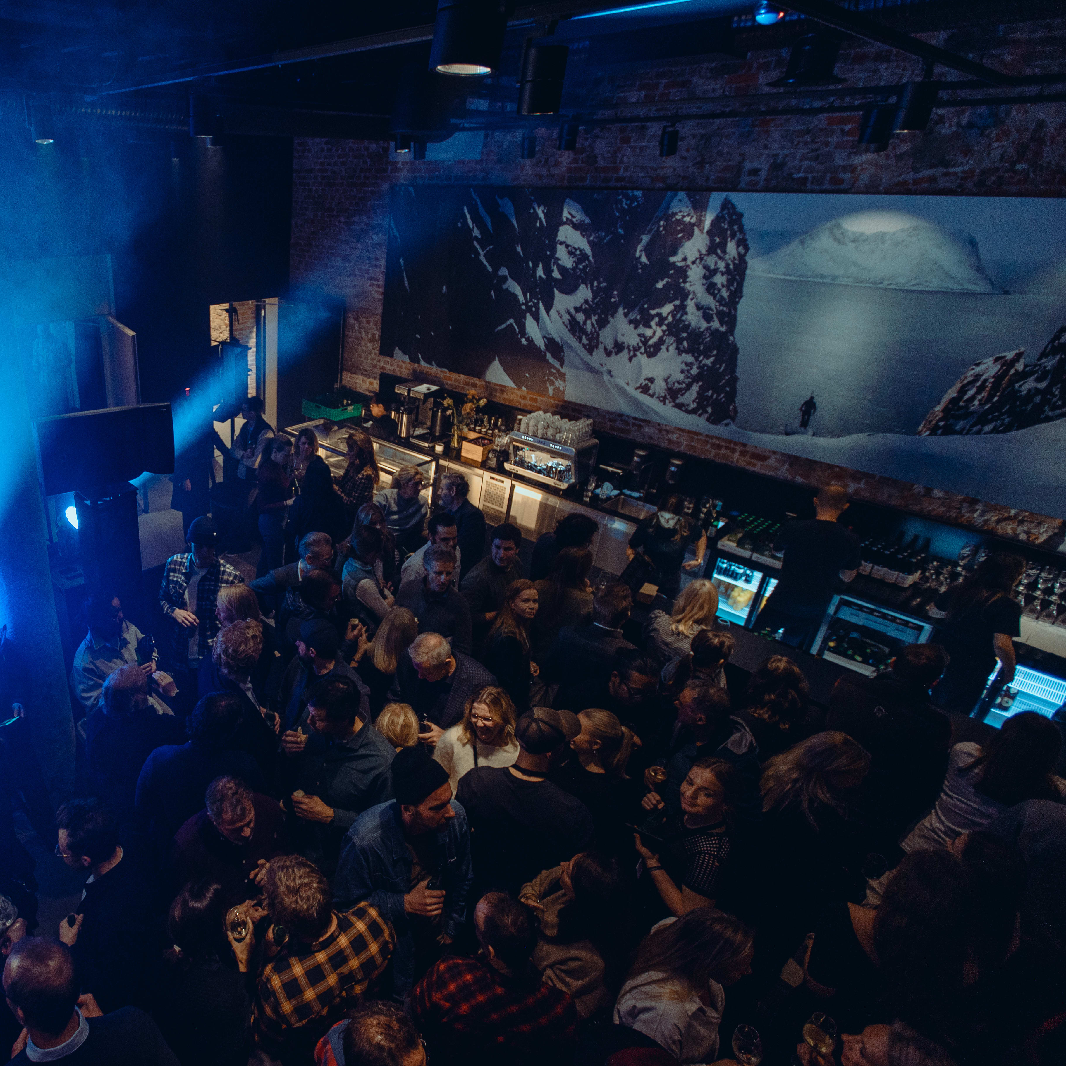 A group of people are standing in a bar. The bar is brightly lit by blue spotlights. There is a bar with a staff member behind it and a large mural of a snowy mountain on the wall behind it. The people are wearing casual clothing, many wearing Norrøna outerwear. 
