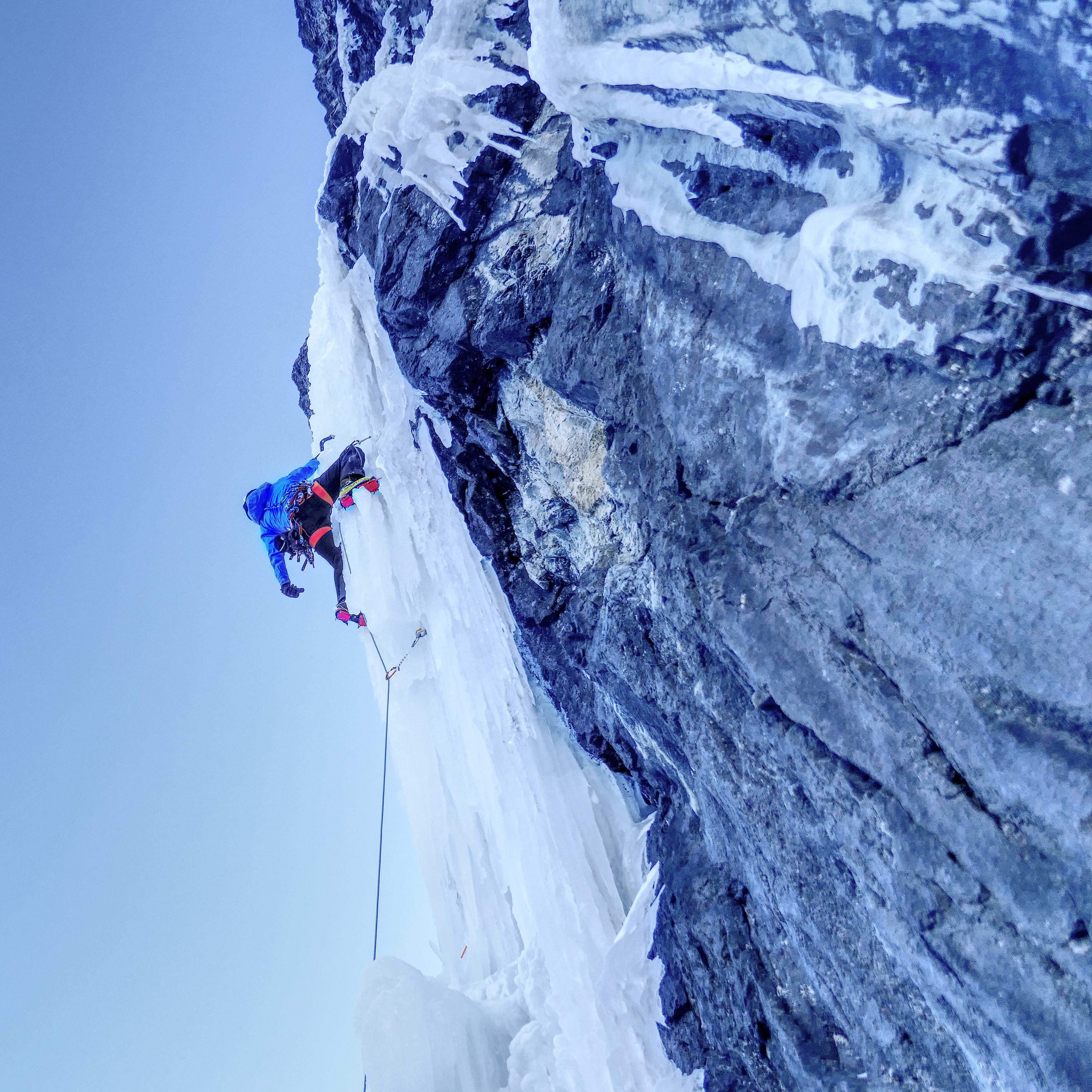 Low angle view of an ice climber wearing a blue Norrøna jacket, ascending a frozen waterfall against a clear blue sky.