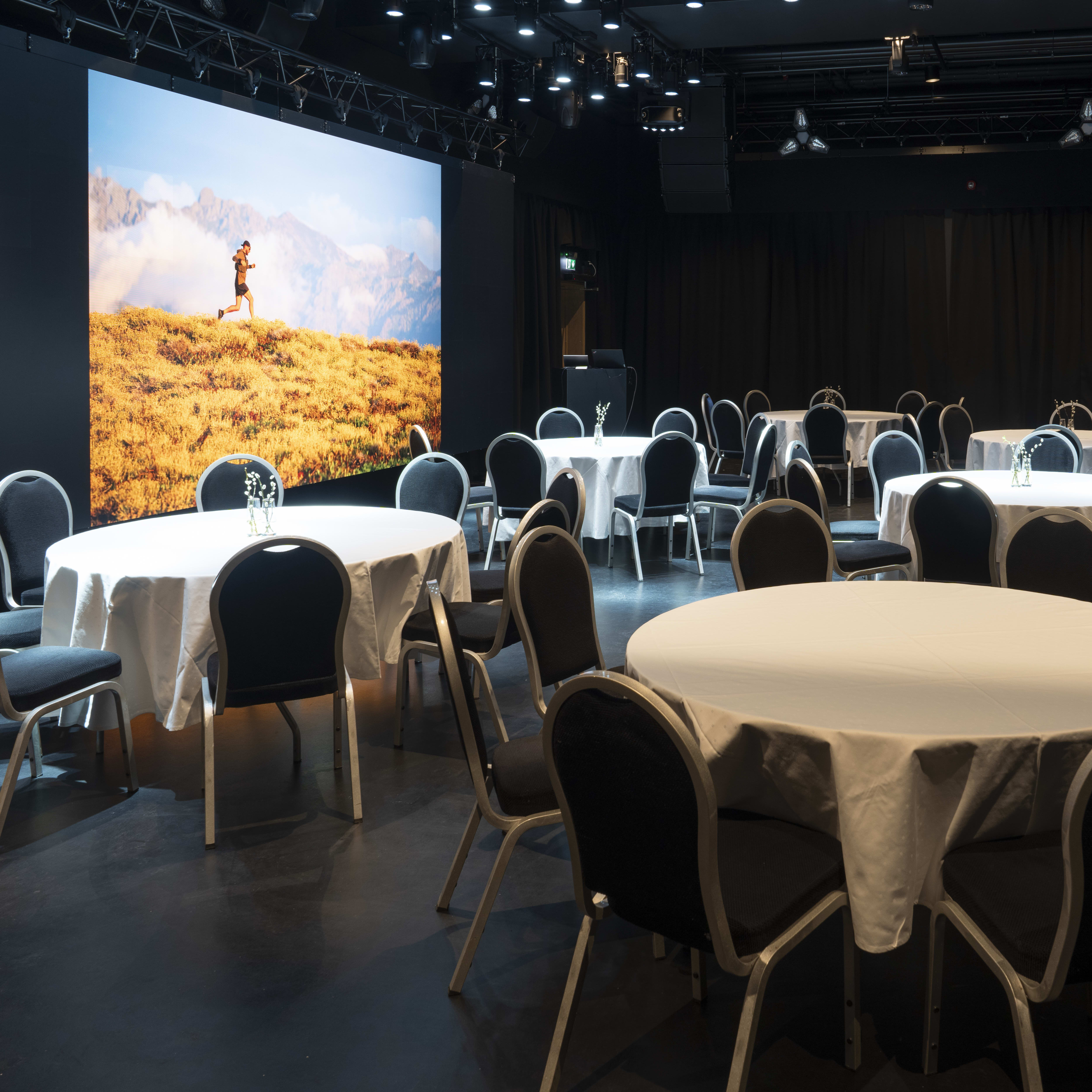 A dark room with round tables and chairs set up for a meeting. A large screen displays a man running through a mountain landscape.