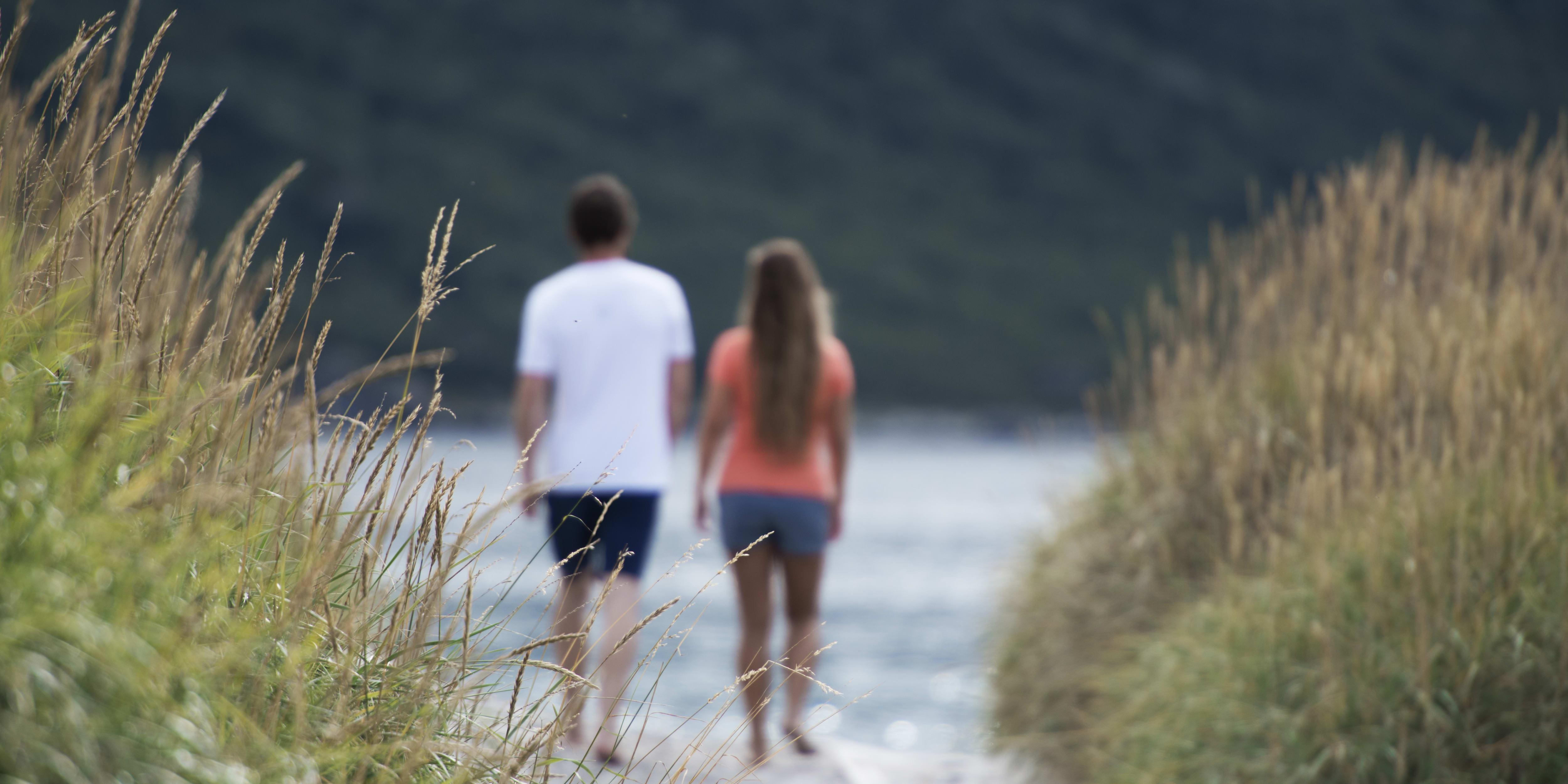 Two people walking on a path through tall grass. The man is wearing a Norrøna shirt. The path leads to a body of water.