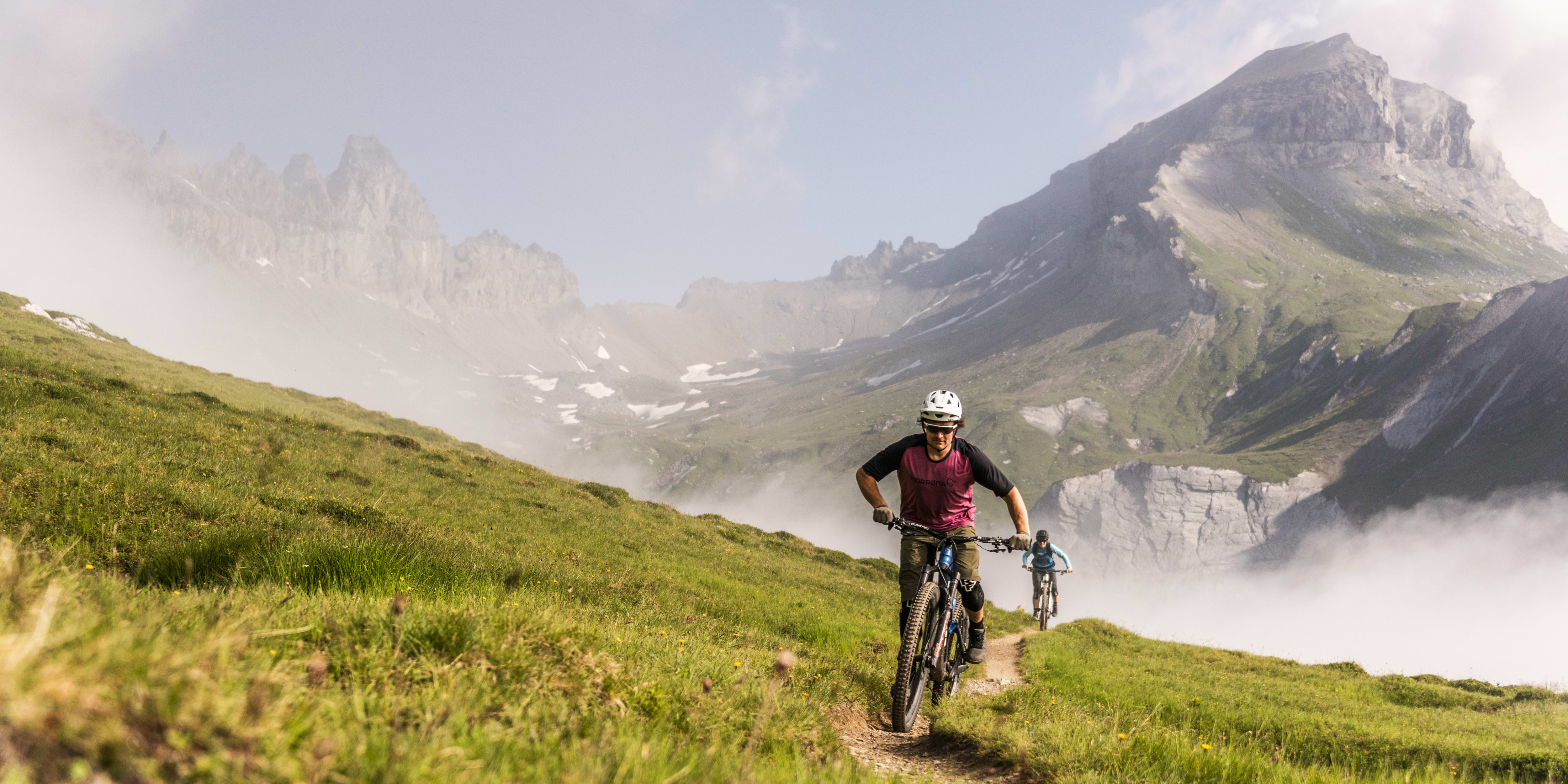 Two mountain bikers are riding up a grassy trail with a mountain in the background. The bikers are wearing helmets and are riding up a steep incline.