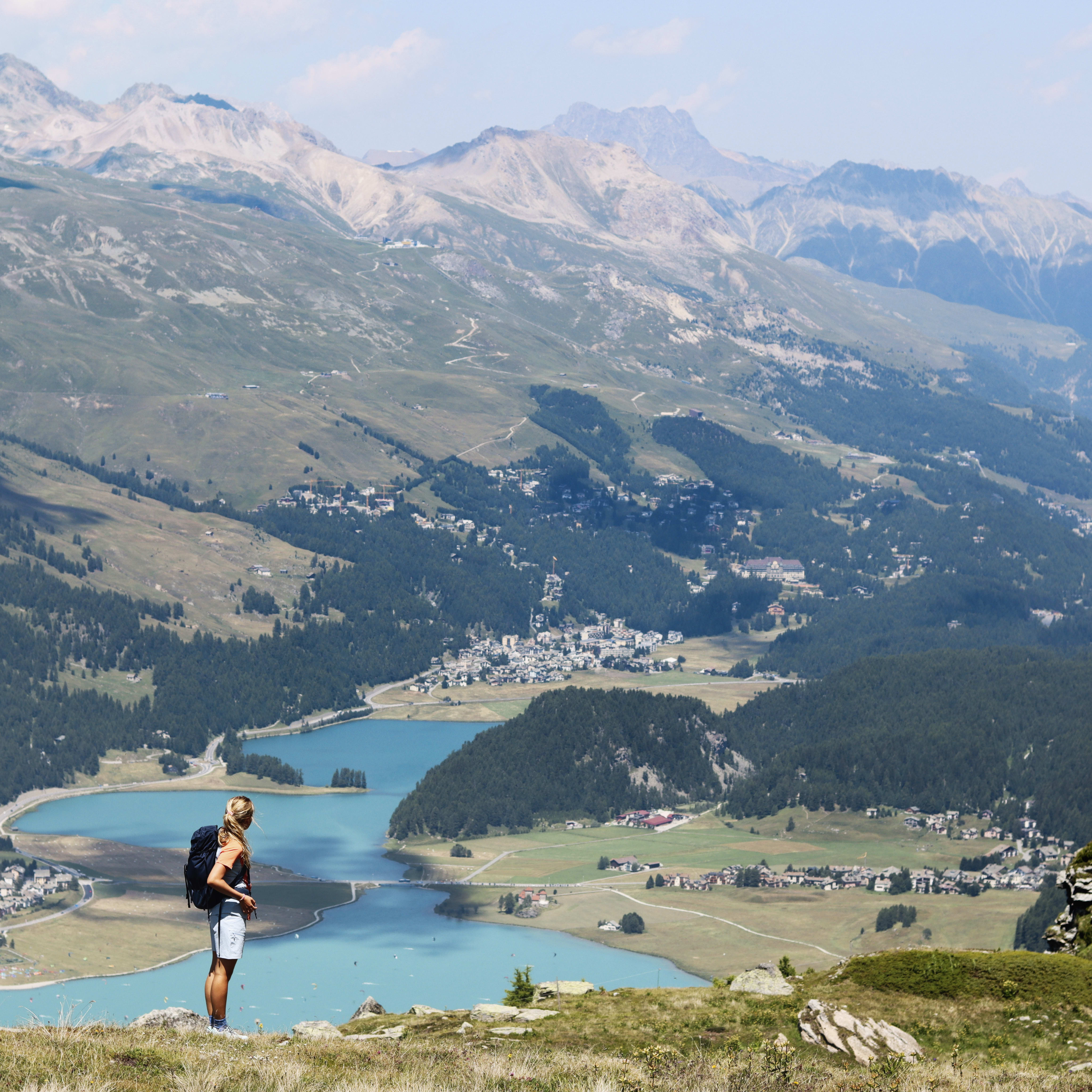 A woman wearing a Norrøna shirt, shorts, and a backpack, stands on a mountaintop overlooking a valley with a lake and a town in the distance. 
