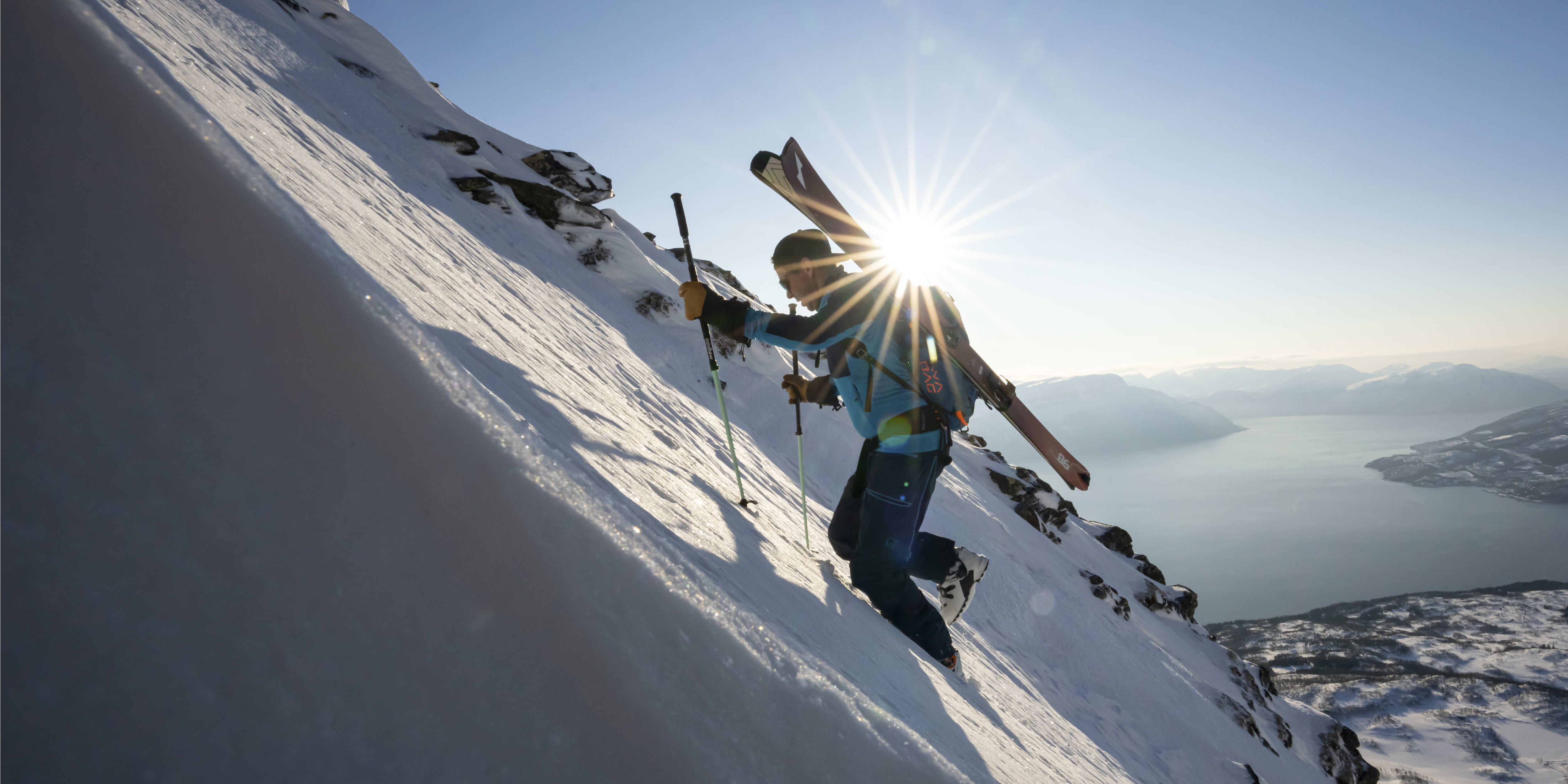 A person wearing Norrøna gear is ski touring uphill in a snow-covered mountain landscape with skis on their back.