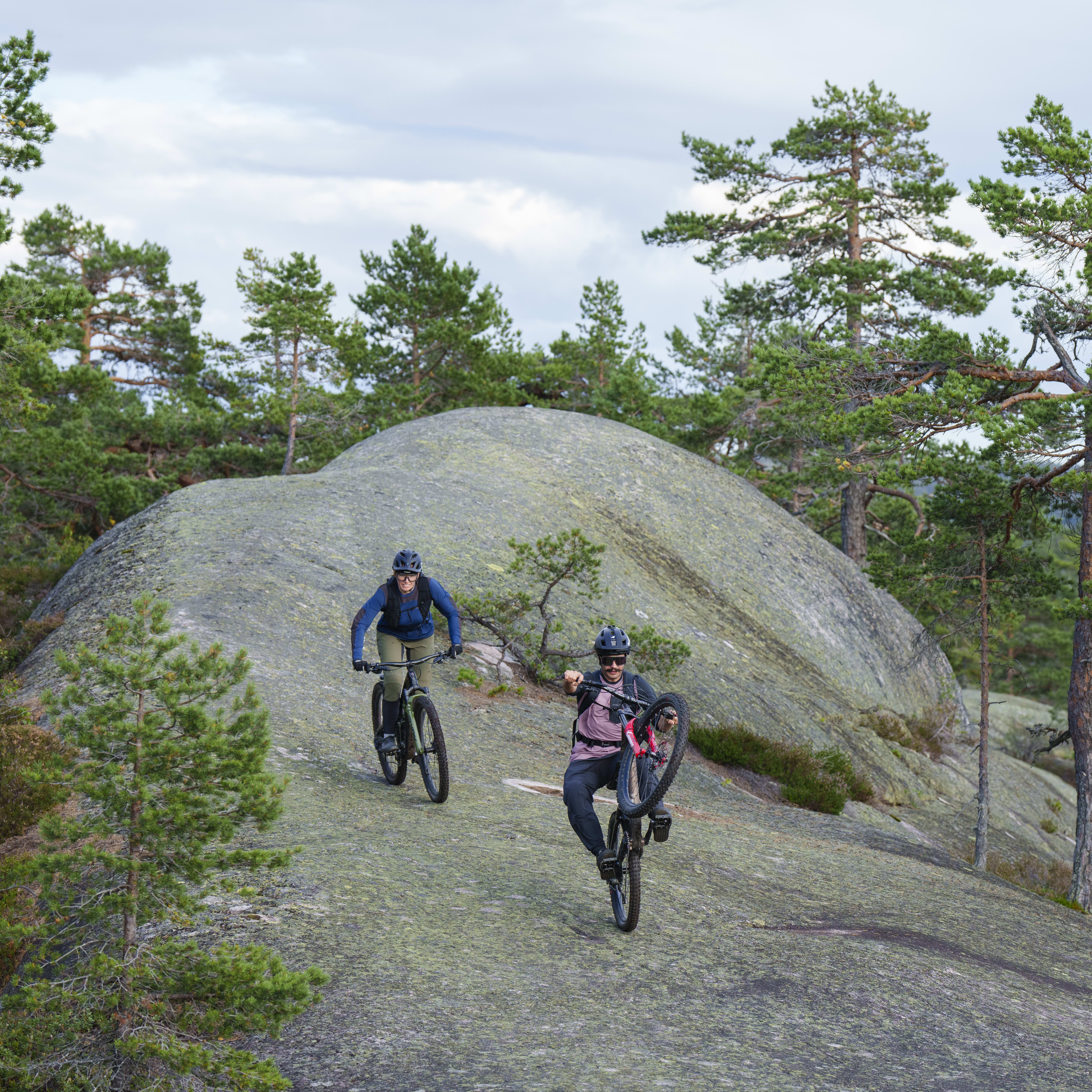Two people are mountain biking on a rocky trail in a forest. The person in the front is doing a wheelie.  Both bikers are wearing Norrøna clothing. 

