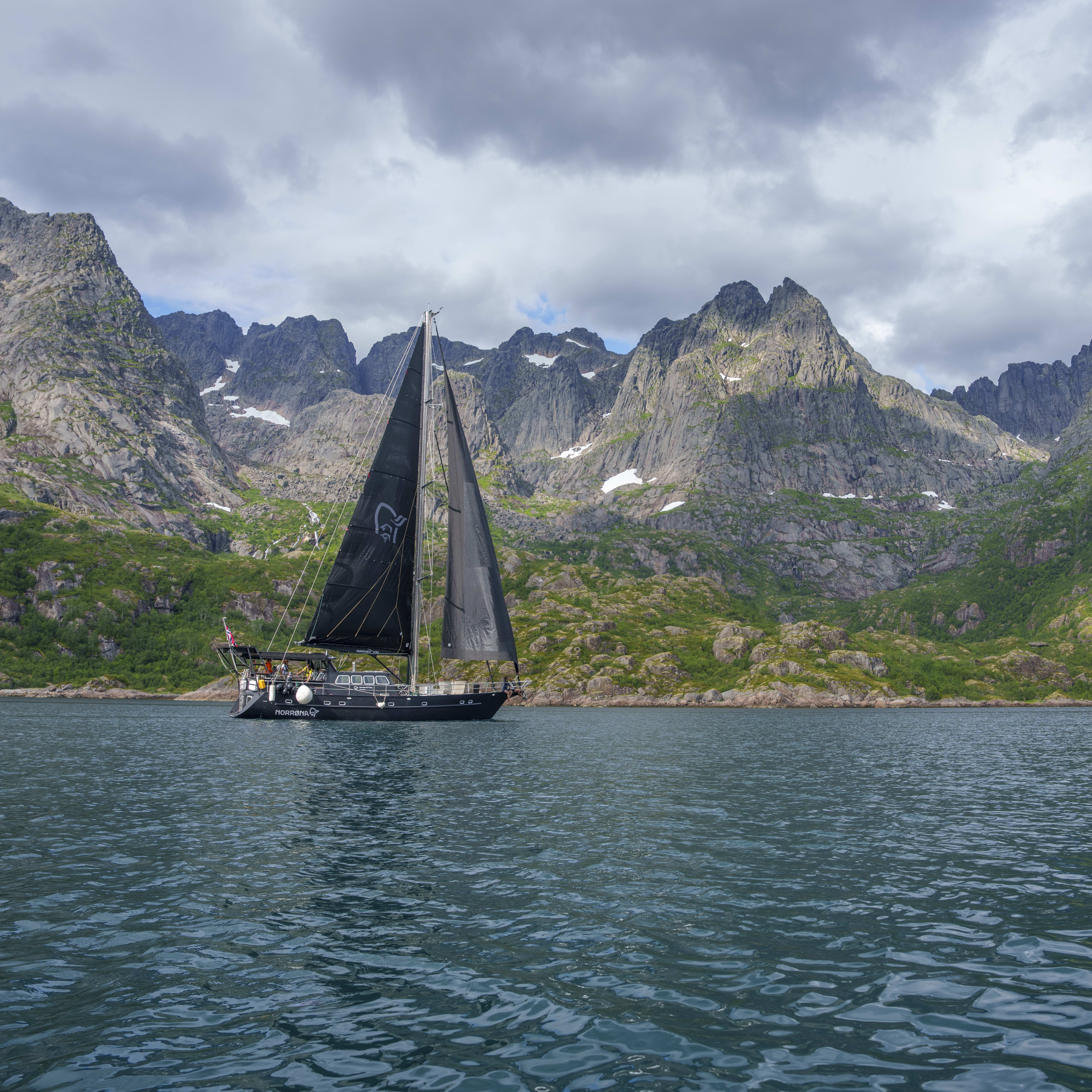 A sailboat with black sails cruises on a body of water beneath jagged, green-covered mountains. "Norrøna" is visible on the side of the boat.