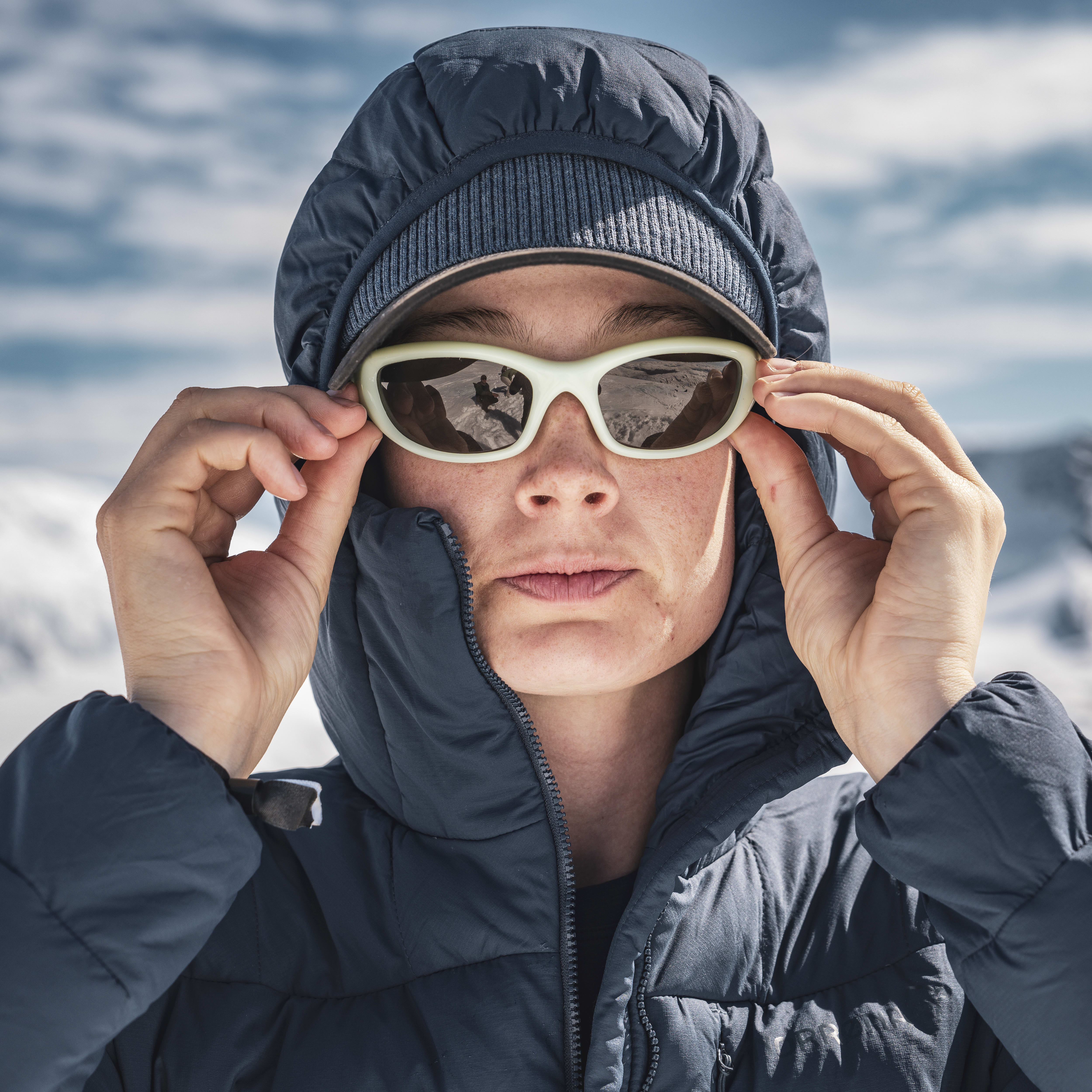 A woman wearing a Norrøna jacket and sunglasses, adjusts her glasses, looking at the camera with a mountain range blurred in the background.