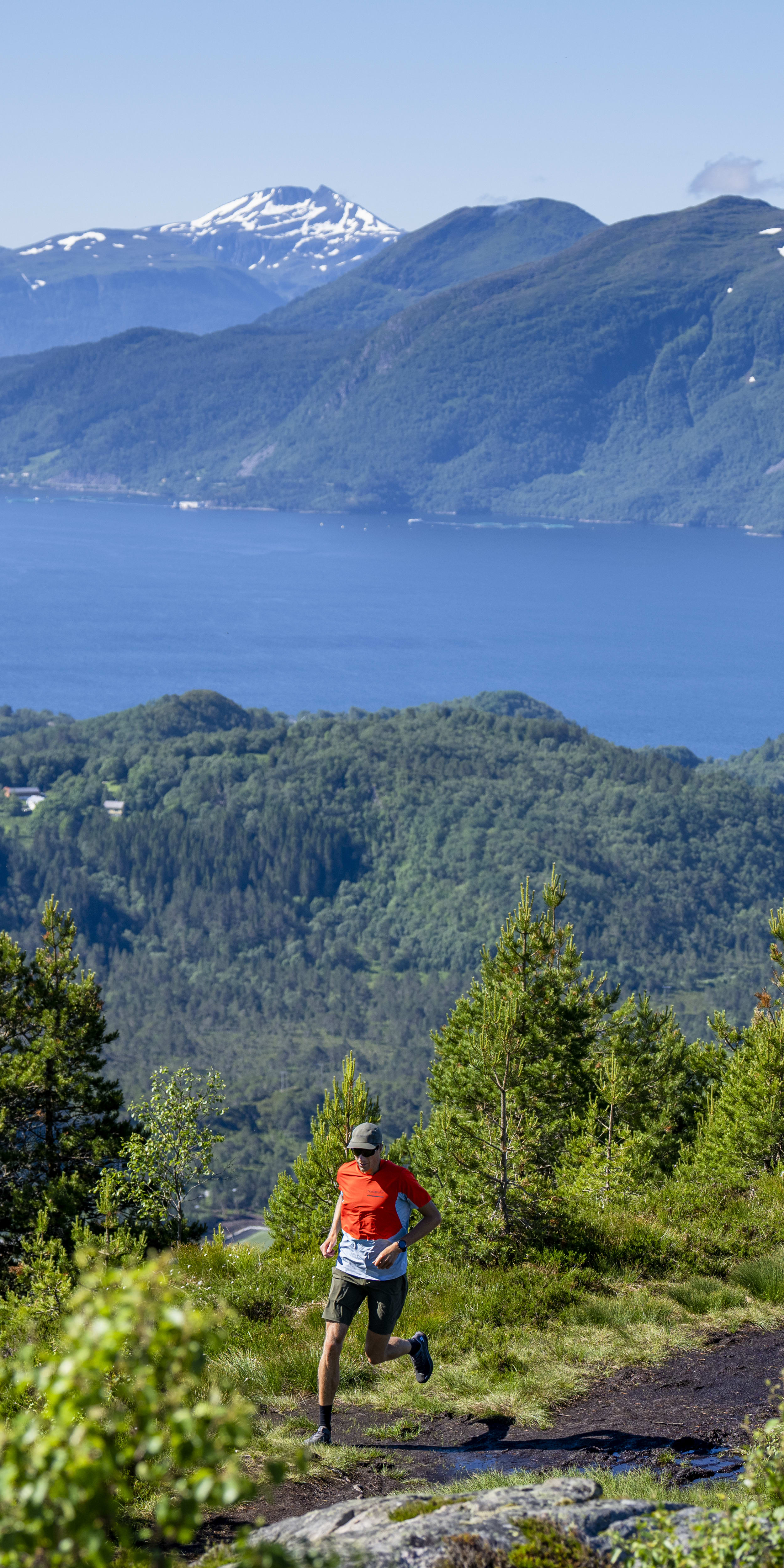 A man wearing Norrøna gear runs along a mountain trail overlooking a fjord.