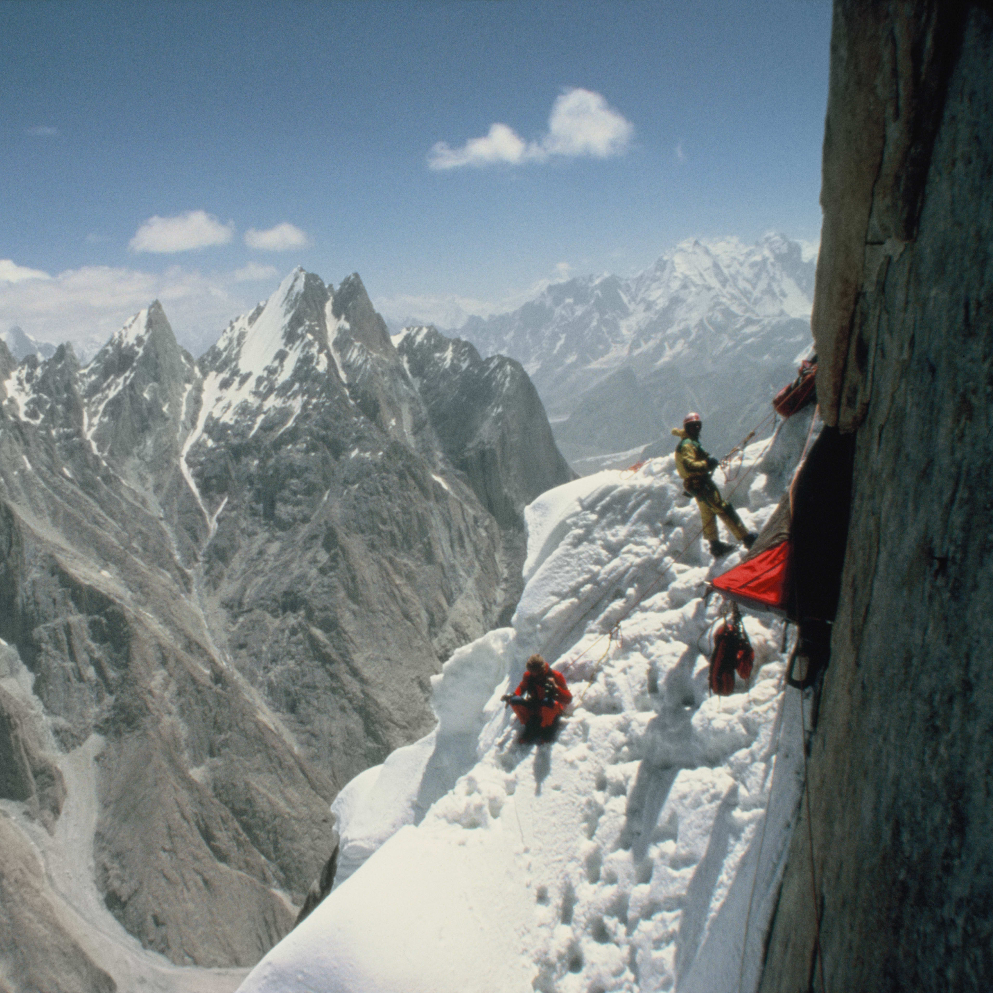 Two climbers on a snowy mountain ridge with a red tent in the background. The view is of a steep, snow-covered slope with a rocky cliff face to the right.  The sky is blue with a few clouds.
