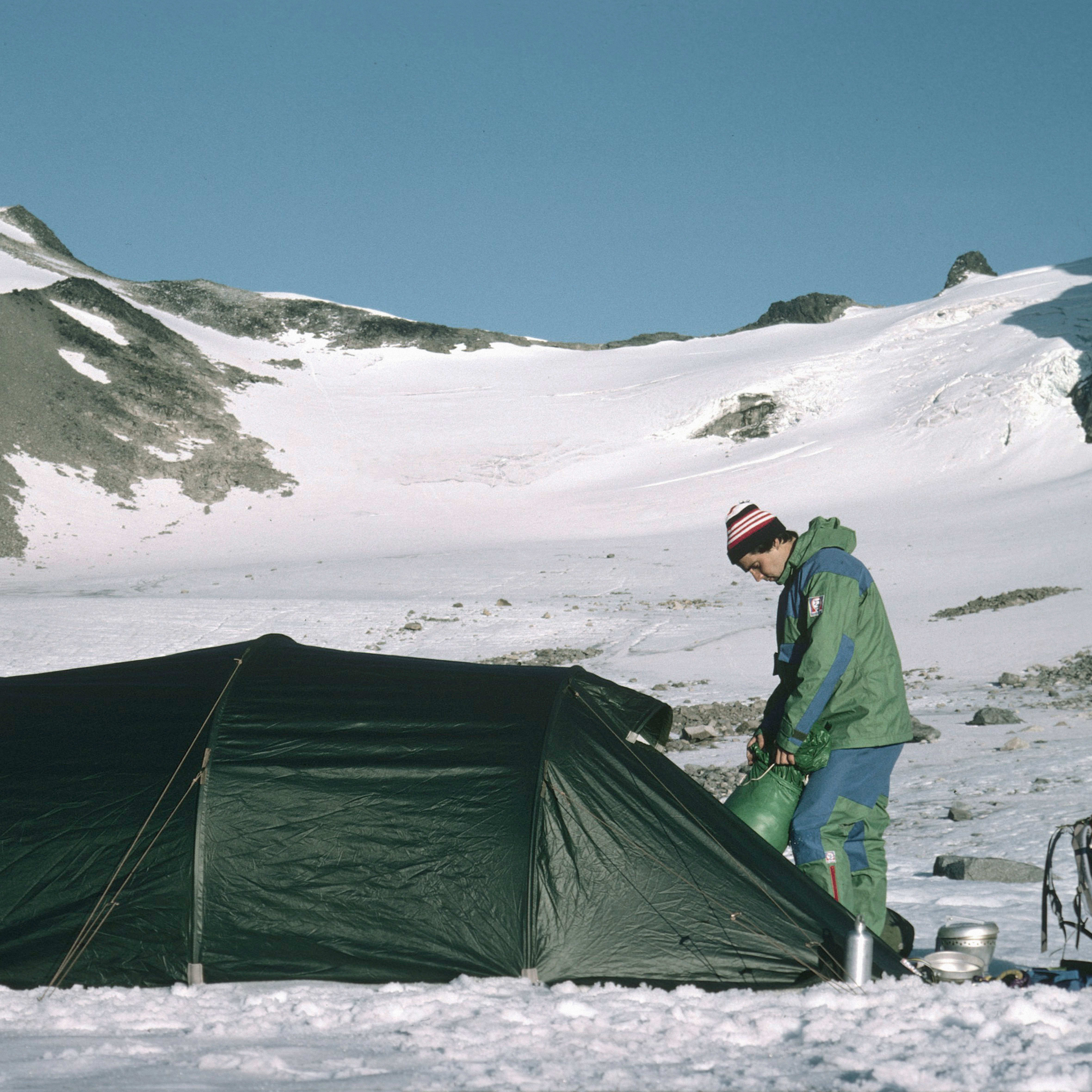 Hiker packing a green bag near a dark green tent. Snowy mountains in the background under a blue sky. The hiker is wearing a Norrøna jacket.
