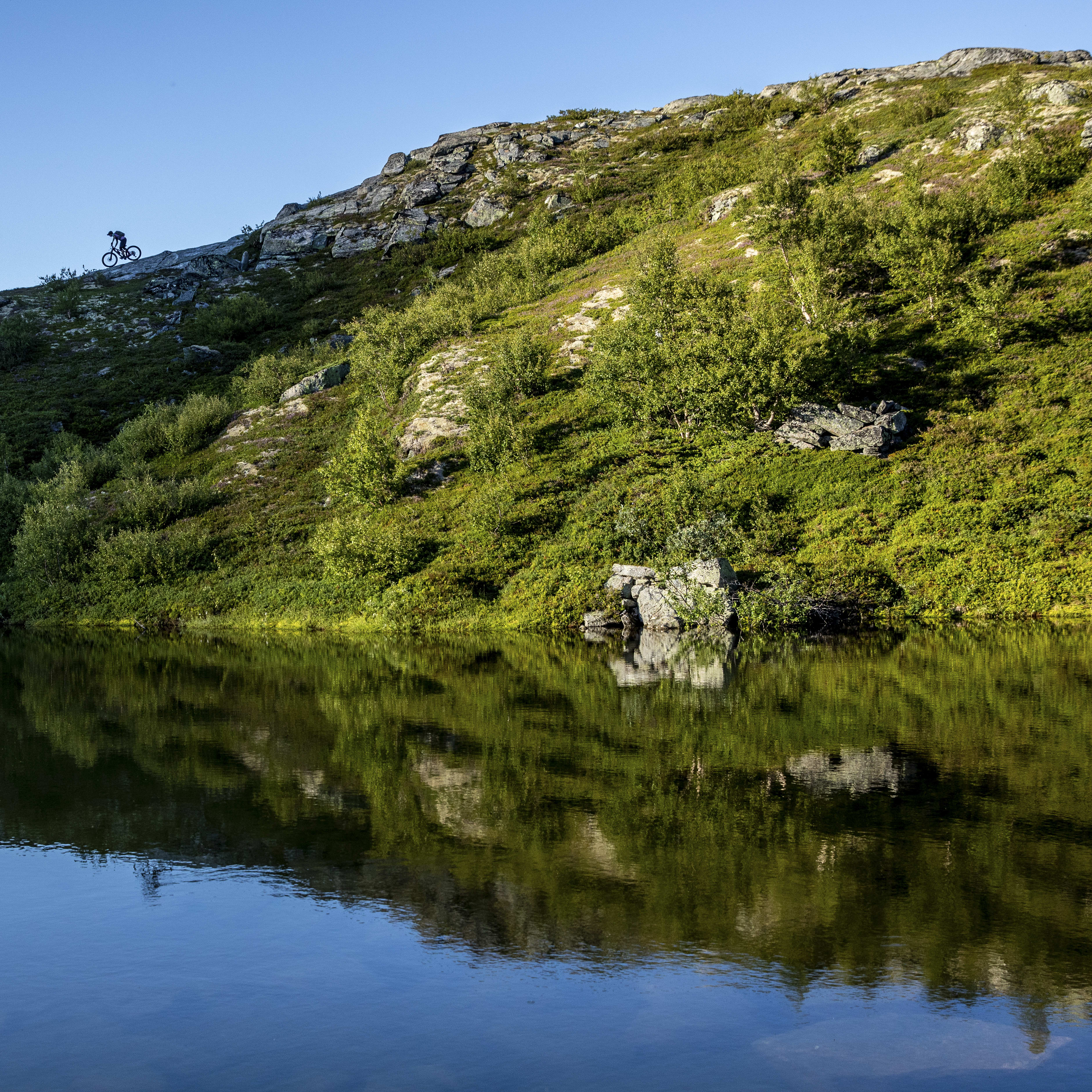 Mountain biker ascends a rocky, green hill reflected in a calm, blue lake under a clear sky.