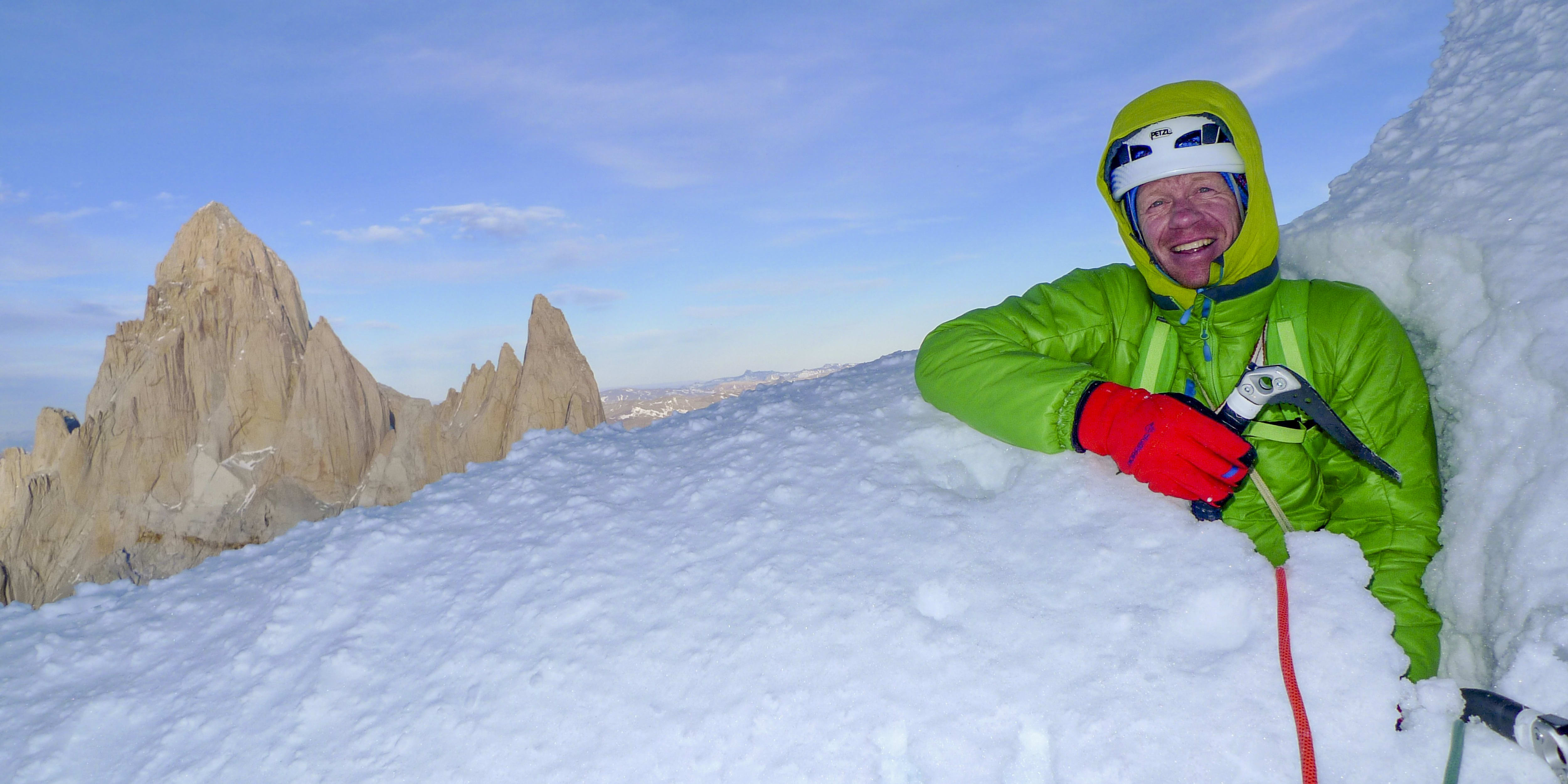A man wearing a green jacket, red gloves, and a helmet is smiling at the camera. He is standing in the snow on a mountain peak, with a large rocky mountain in the background. Two ice axes and a red rope are in the snow around the man.
