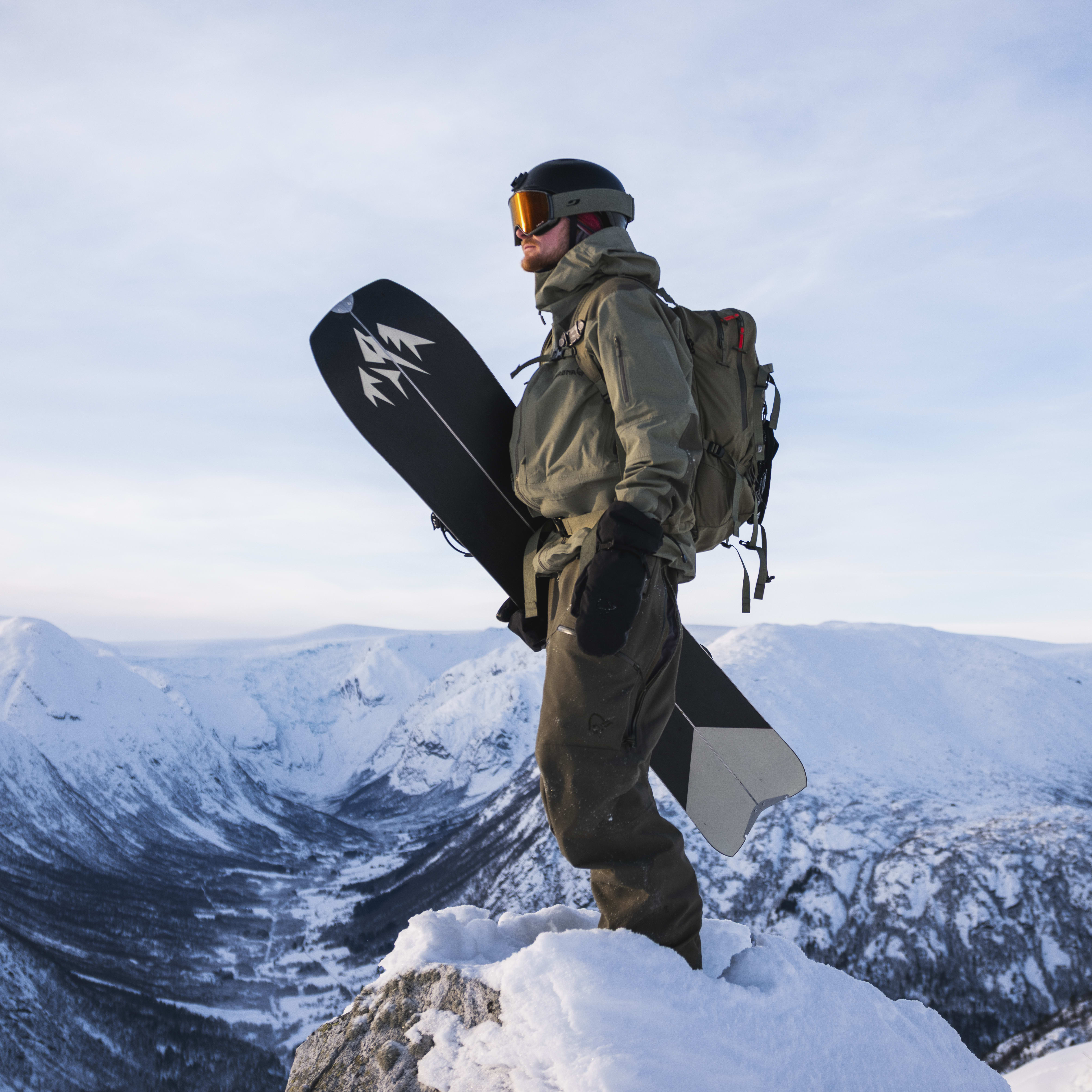 A snowboarder with a Norrøna jacket and pants stands on a snowy peak holding a snowboard. Snowy mountains and blue sky in background.