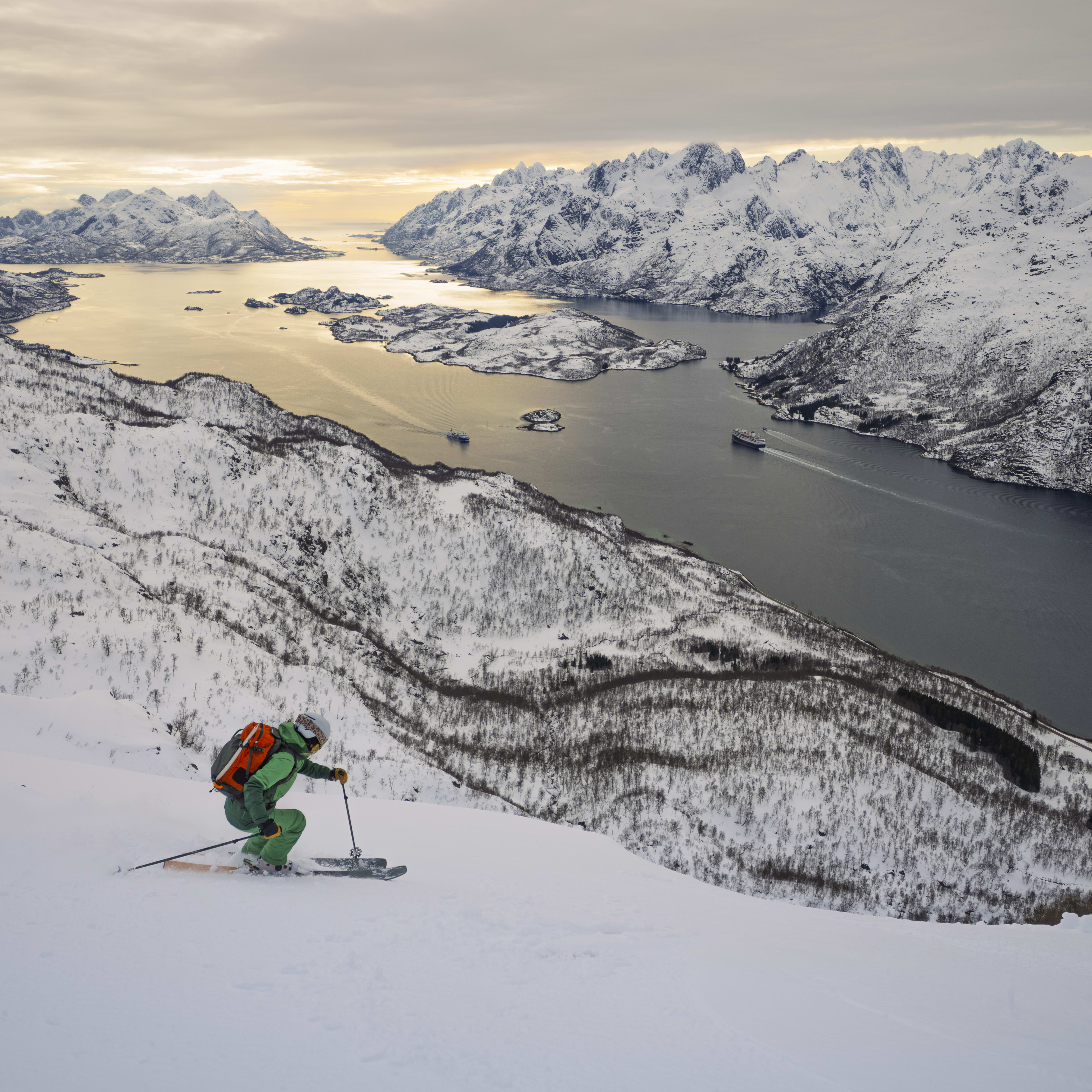 A skier wearing a Norrøna jacket and backpack descends a snowy slope, overlooking a fjord surrounded by snow-capped mountains. 
