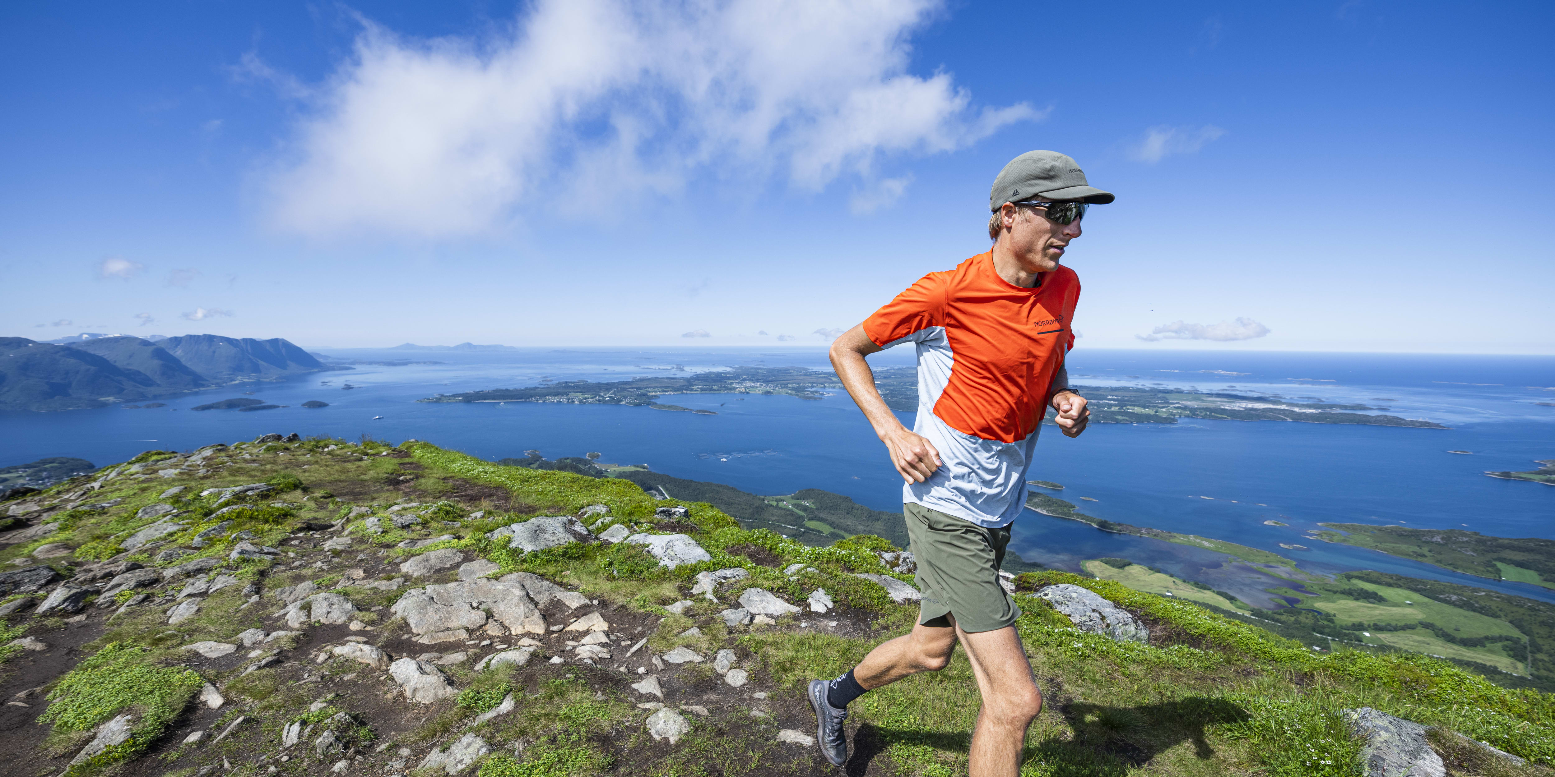A man in a Norrøna shirt and shorts runs along a rocky mountain path overlooking a scenic fjord. 
