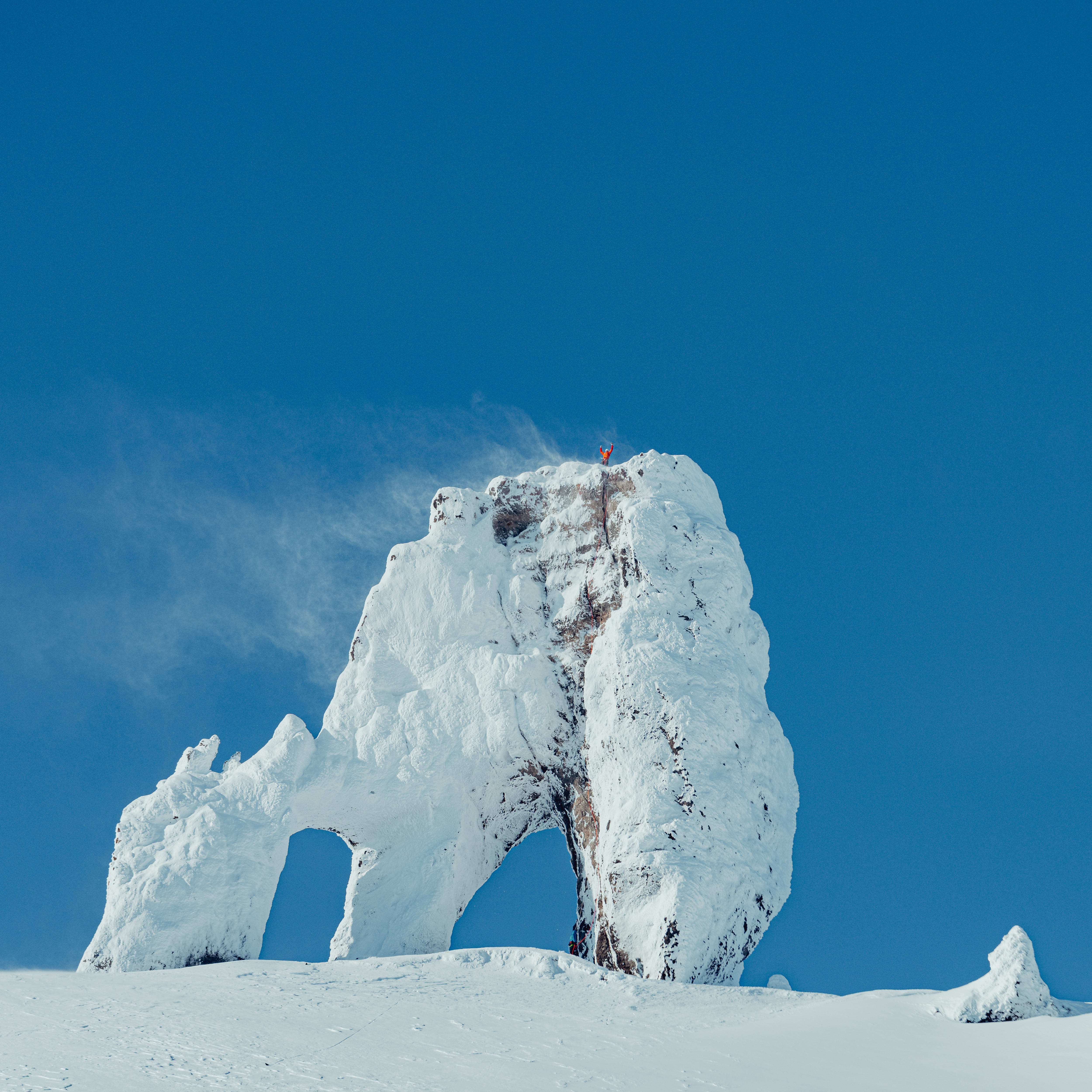 A person wearing a Norrøna jacket stands on top of a snow-covered rock formation against a clear blue sky. 
