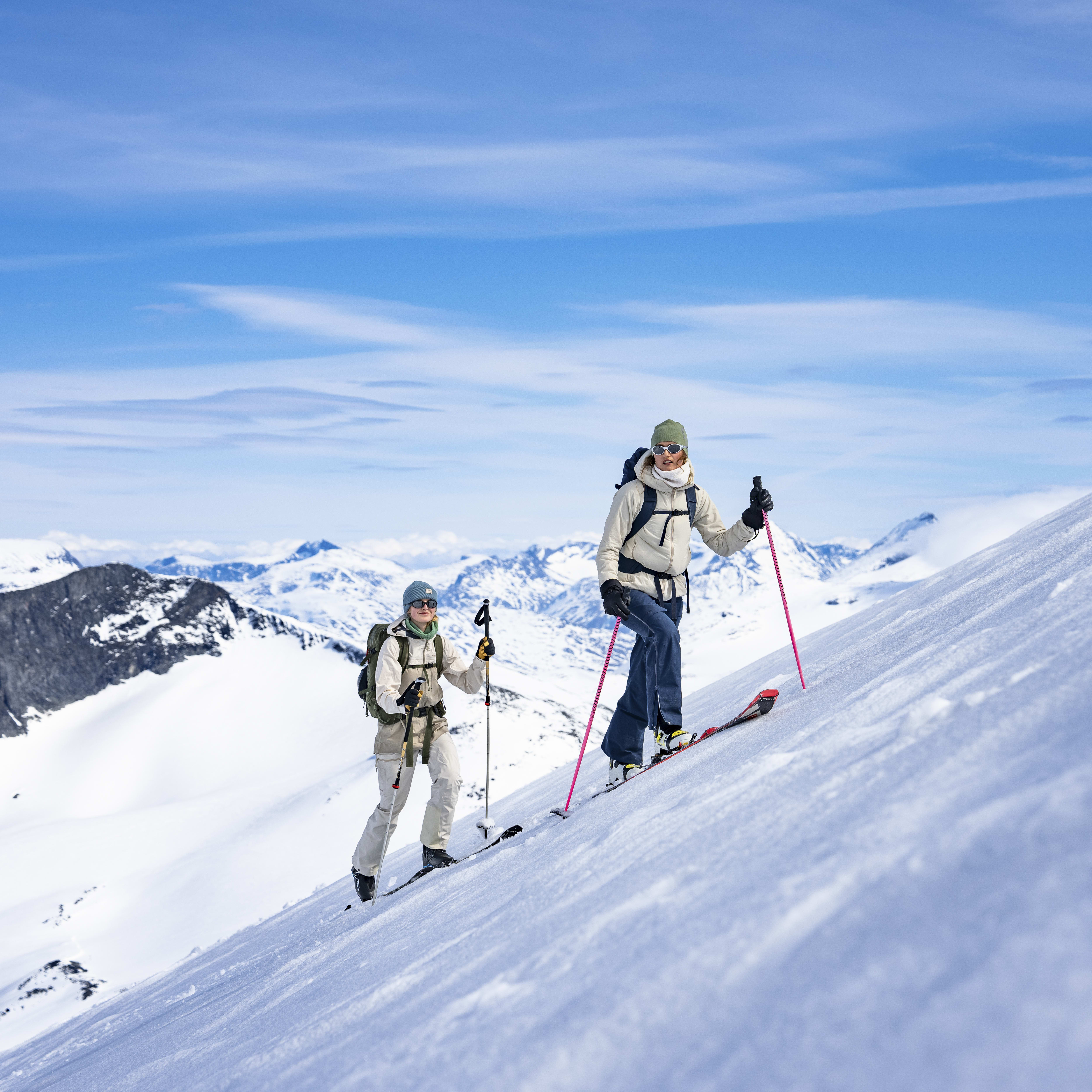 Two people wearing Norrøna gear ski across a snow covered mountain range under a bright blue sky with white clouds. 

