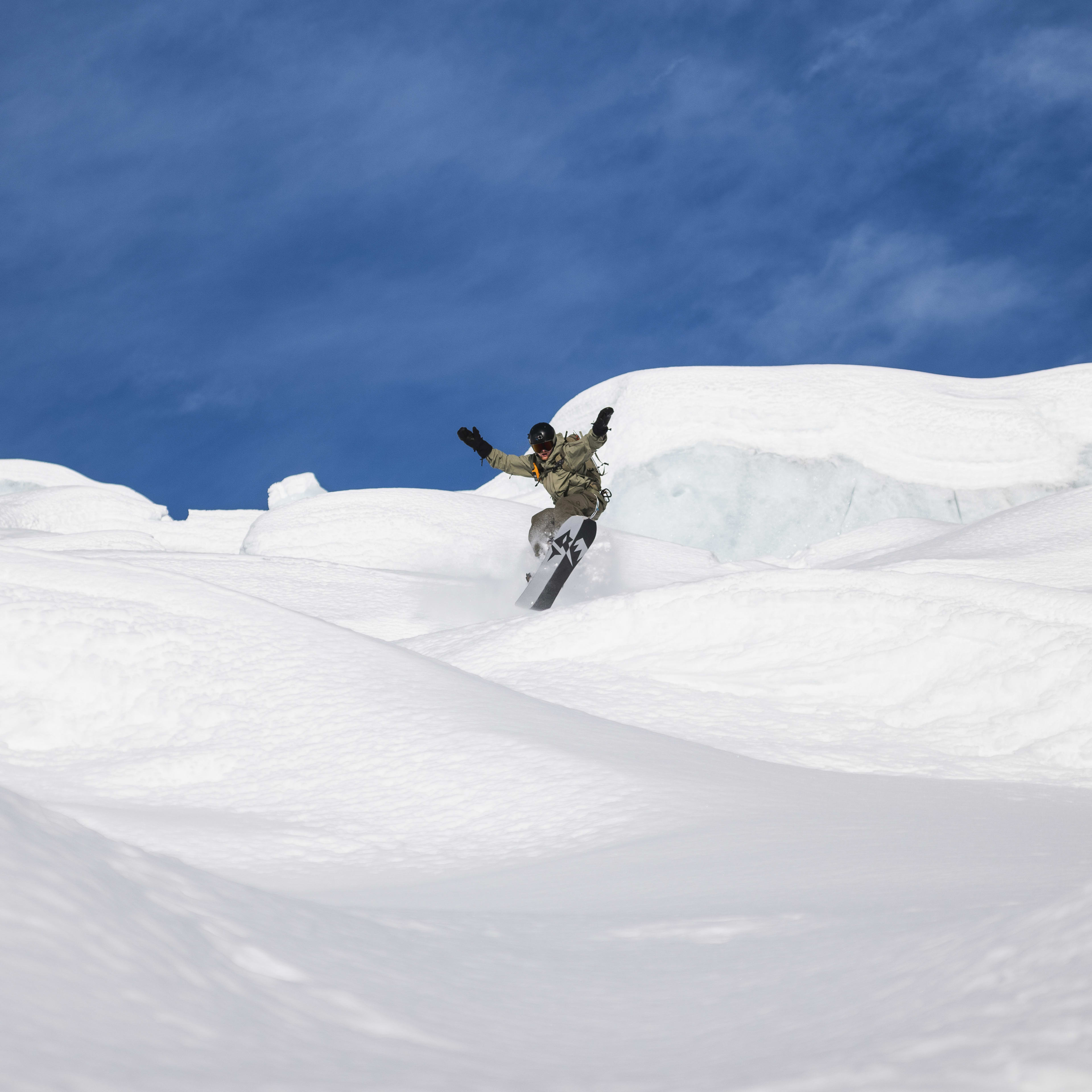 A snowboarder in a Norrøna jacket rides down a snowy slope under a bright blue sky.