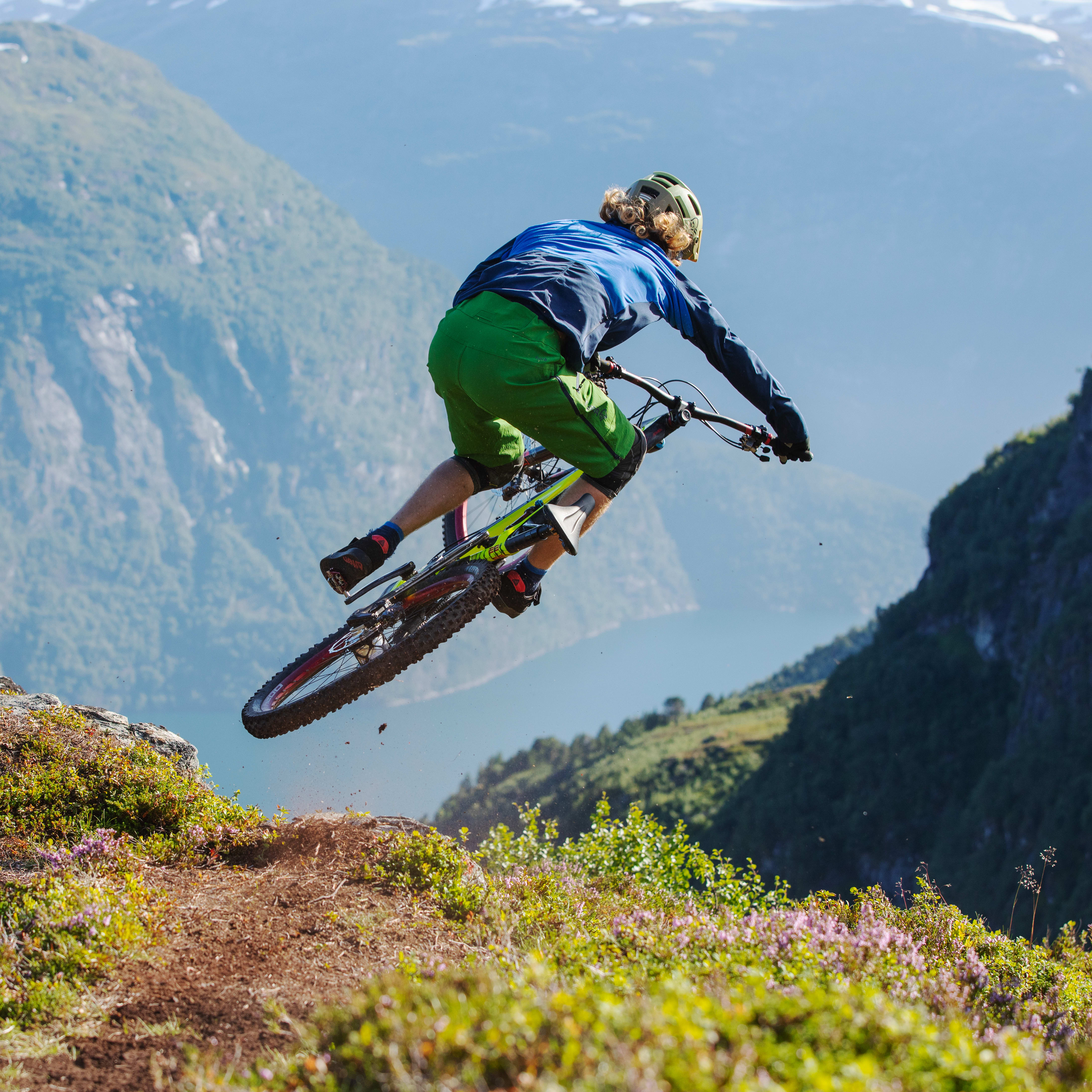 A person wearing a Norrøna outfit jumps off a cliff on a mountain bike with a stunning view of the mountains in the background.