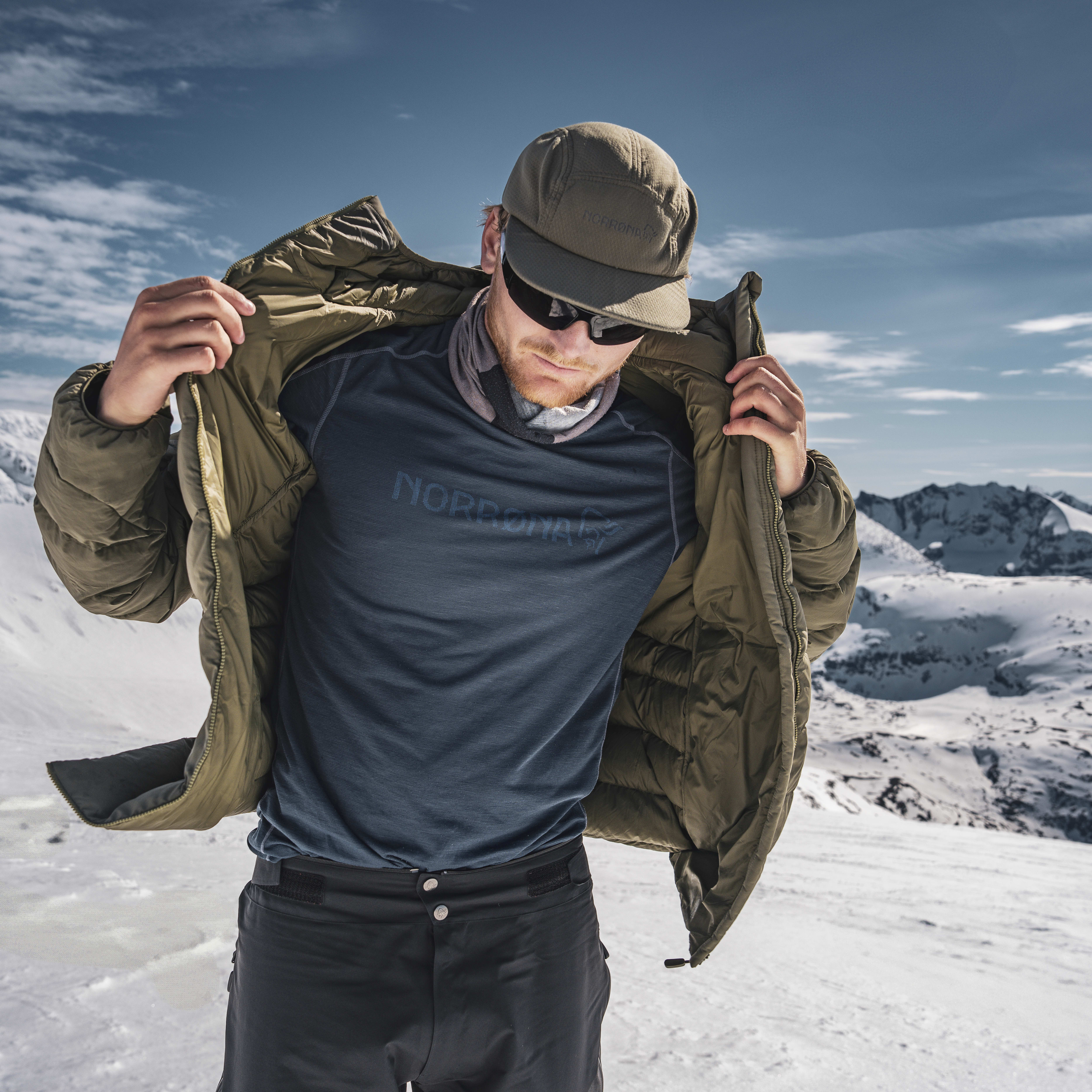 A man wearing a Norrøna jacket and sunglasses stands in snowy mountains. 
