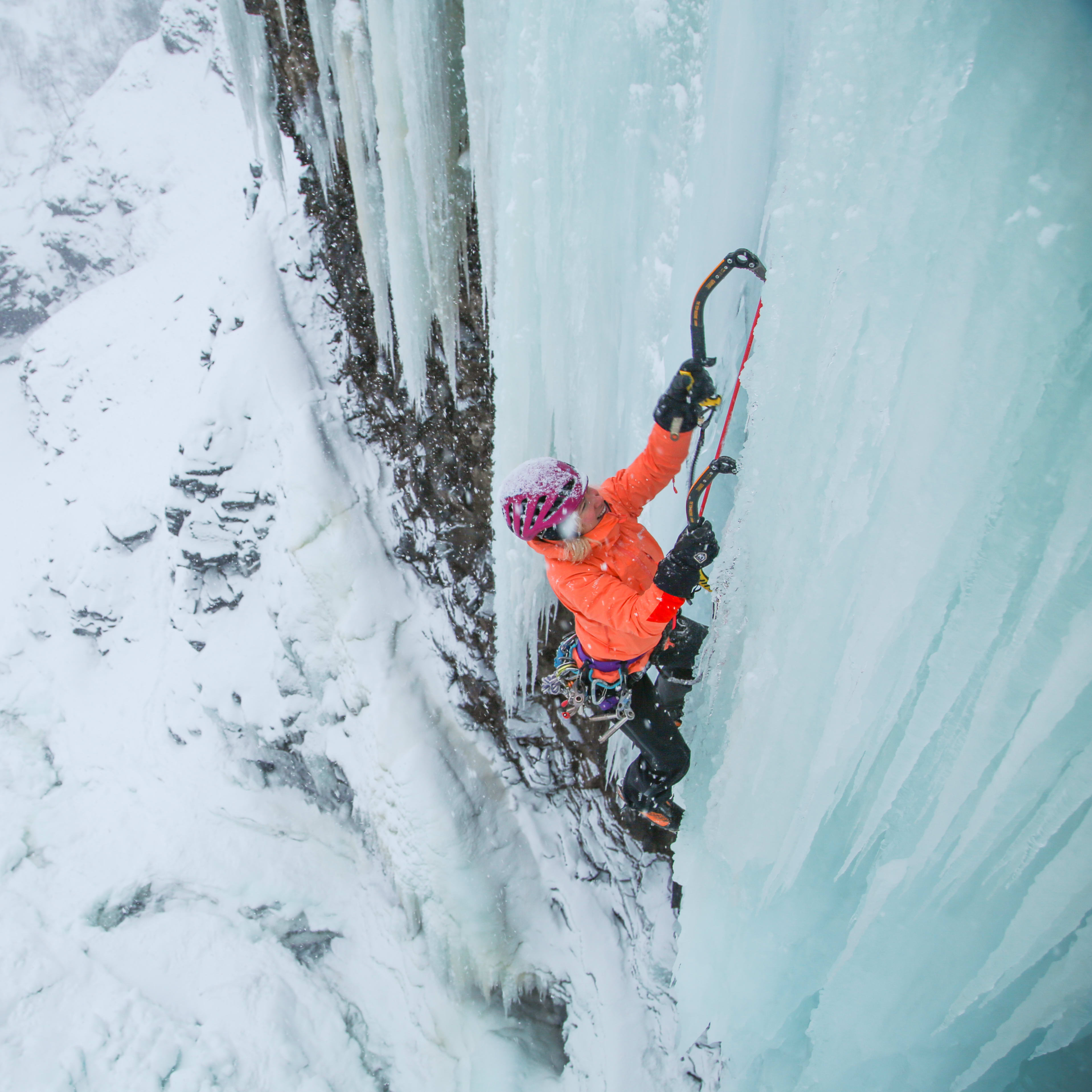 A climber wearing a Norrøna jacket and helmet ascends an ice wall.  
