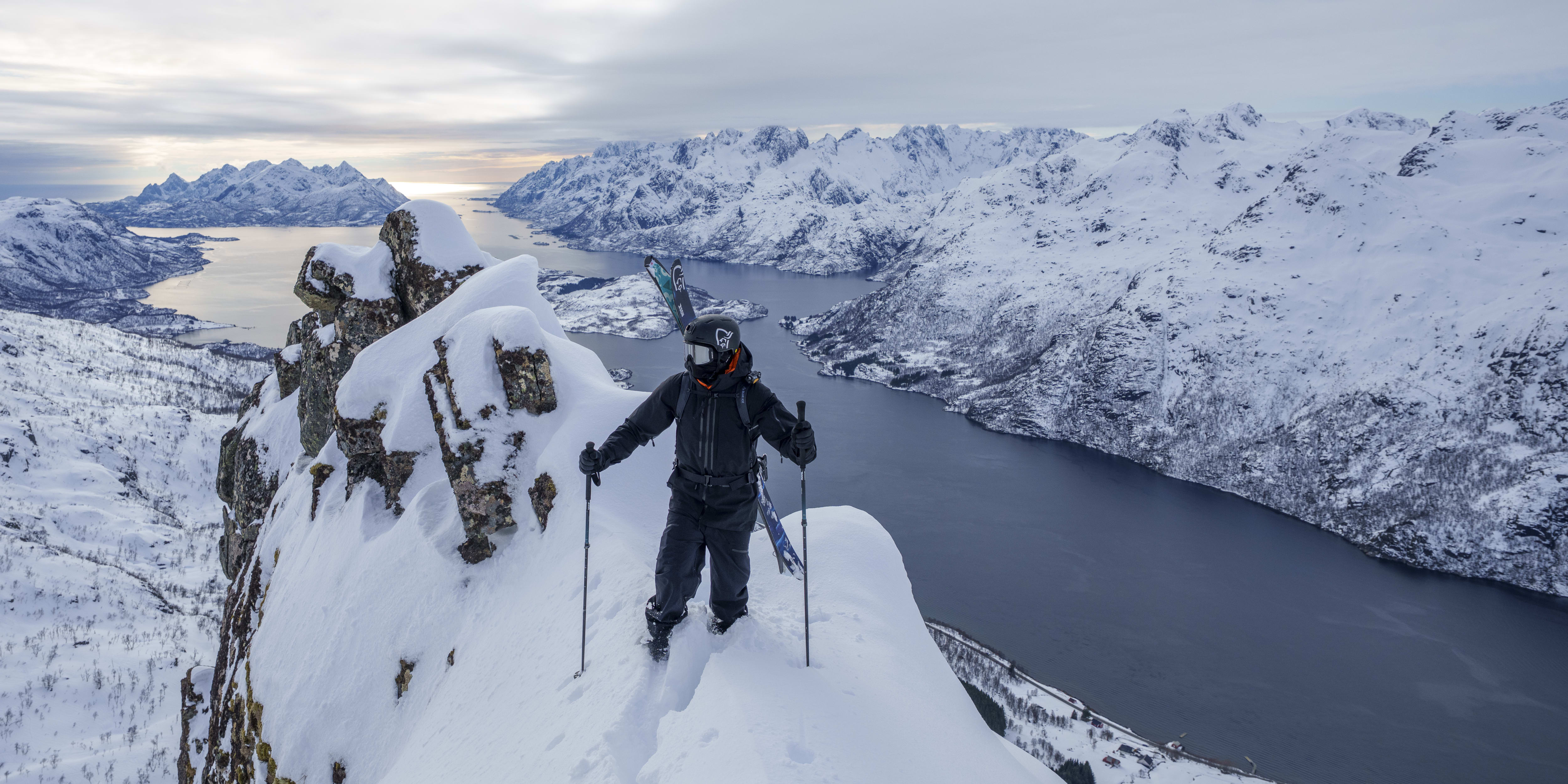 A skier wearing Norrøna clothing stands on a snowy mountaintop, looking out over a fjord.