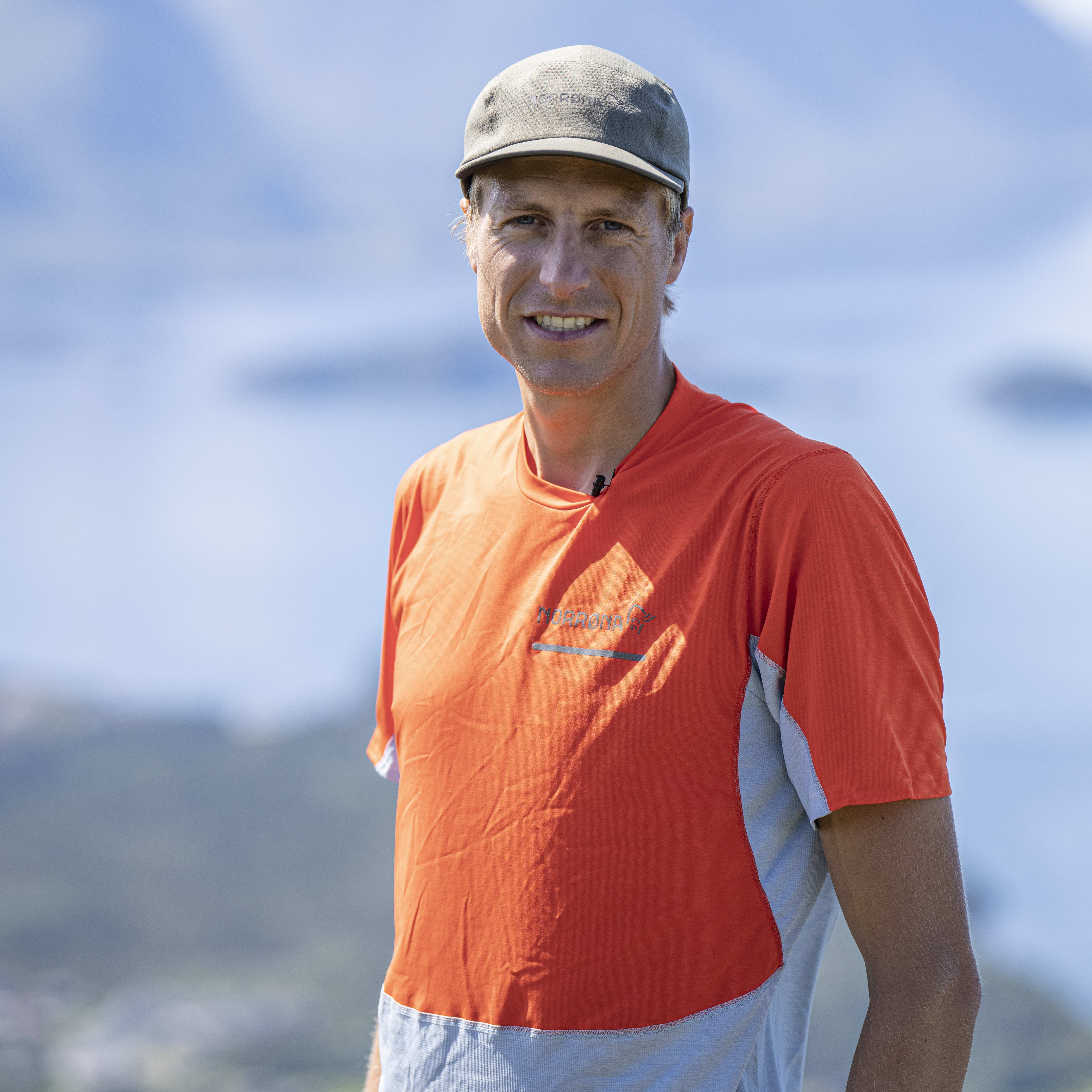 A man smiling at the camera. He's wearing a Norrøna shirt and a hat, standing in front of a blurred mountain background.