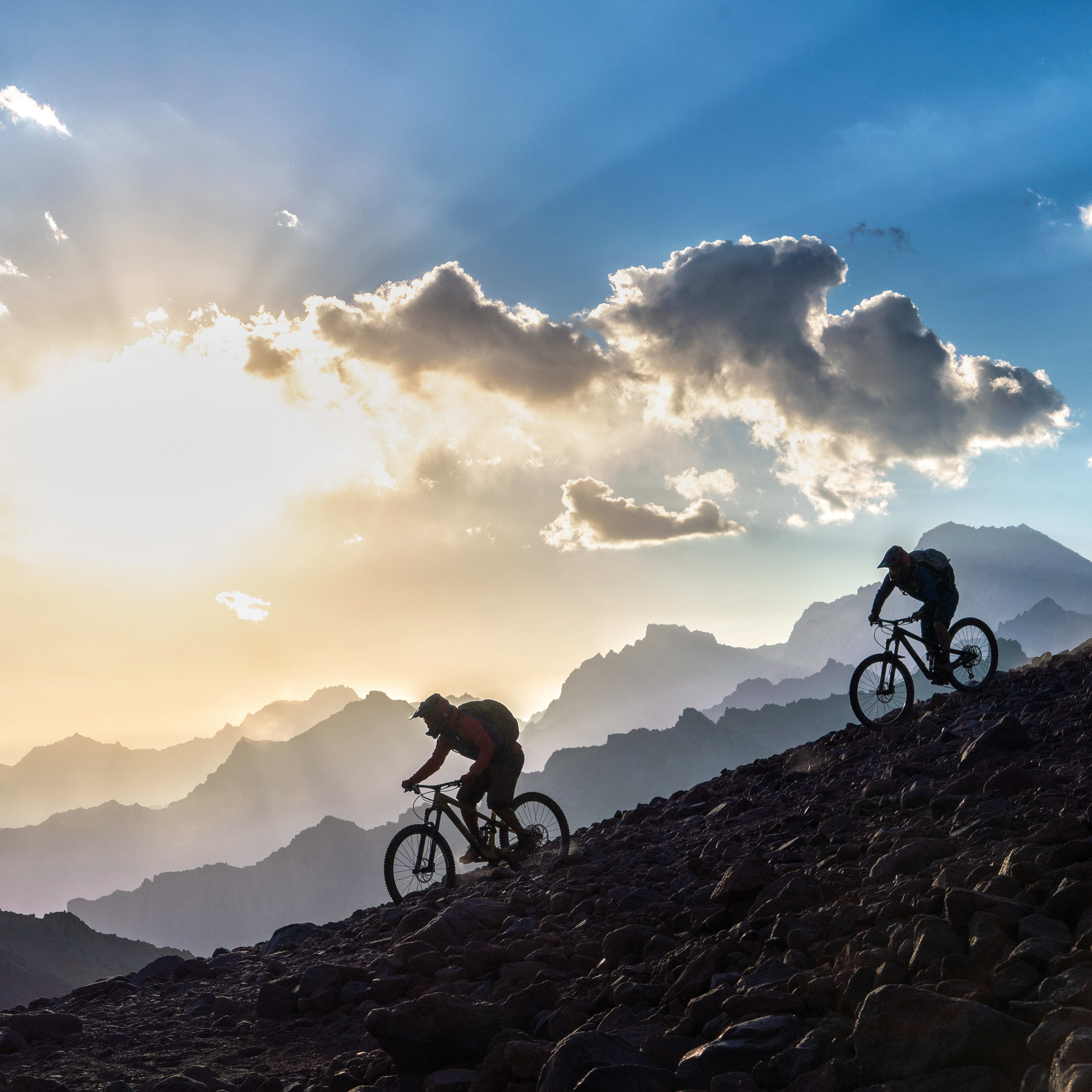 Two mountain bikers riding Norrøna gear down a rocky path with a mountain range and a sunset in the background. 
