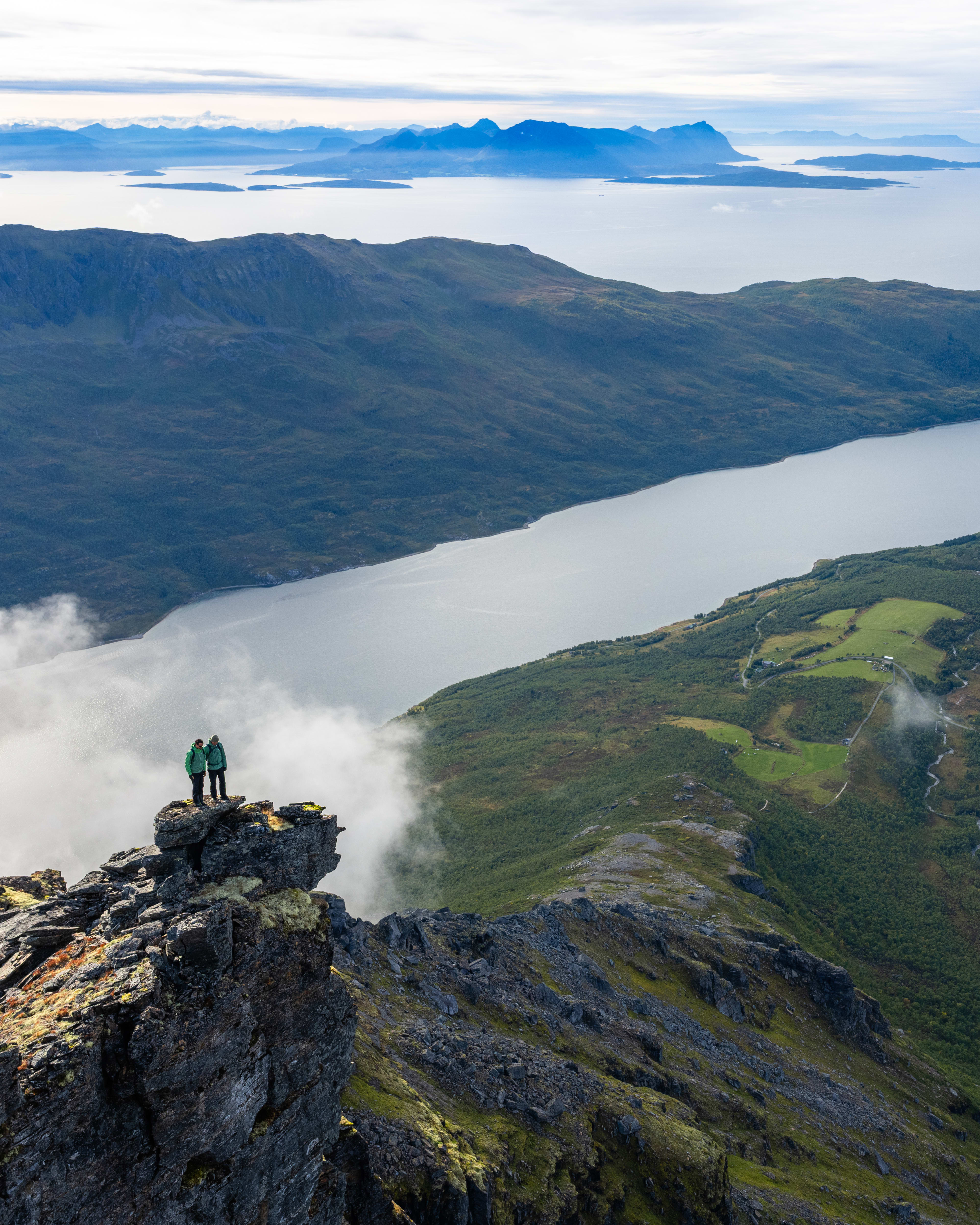 Two hikers in Norrøna gear stand on a rocky summit overlooking a vast fjord landscape.