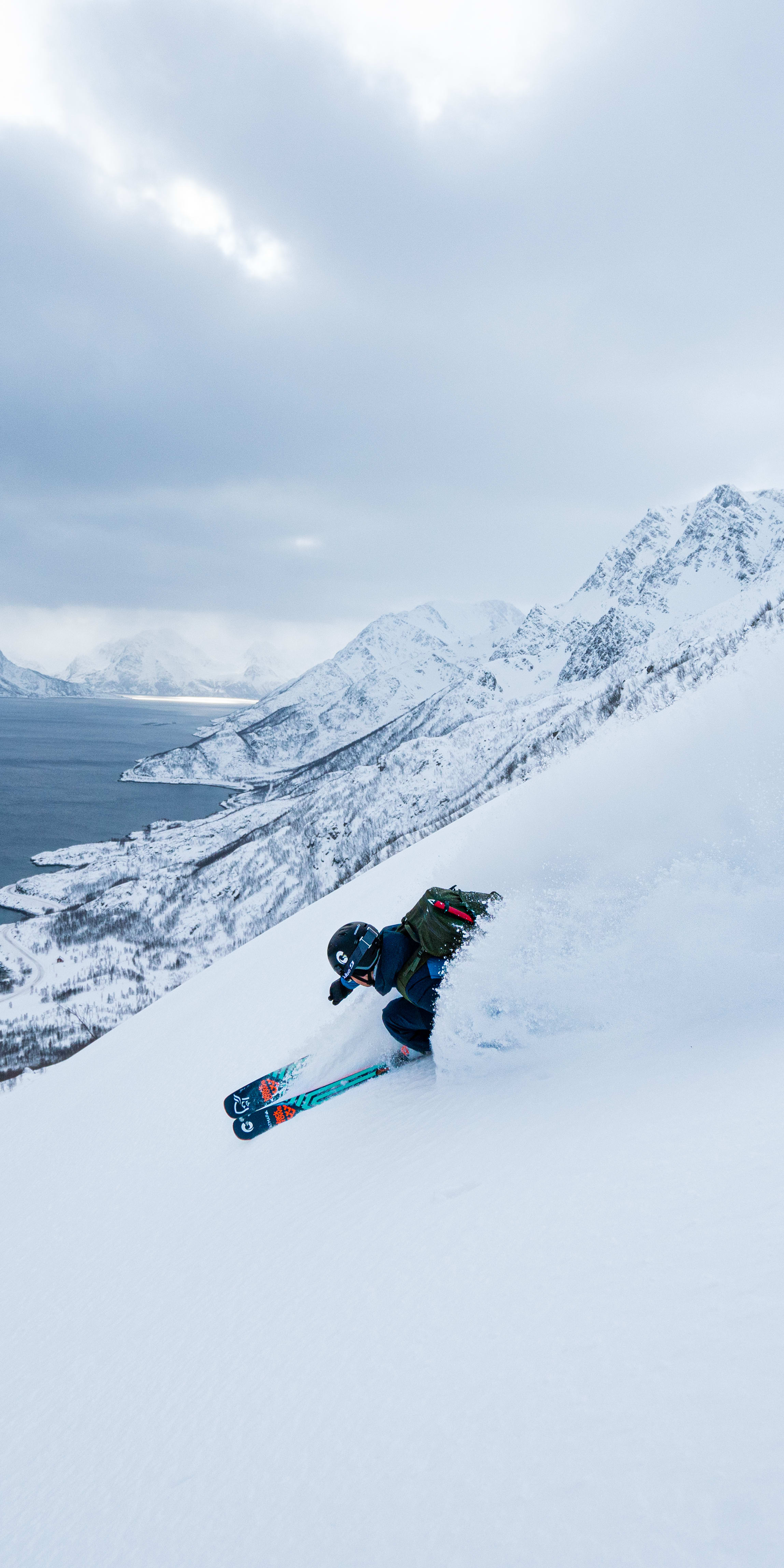 A skier wearing Norrøna gear descends a snowy mountain slope, with a fjord and distant mountains in the background. 
