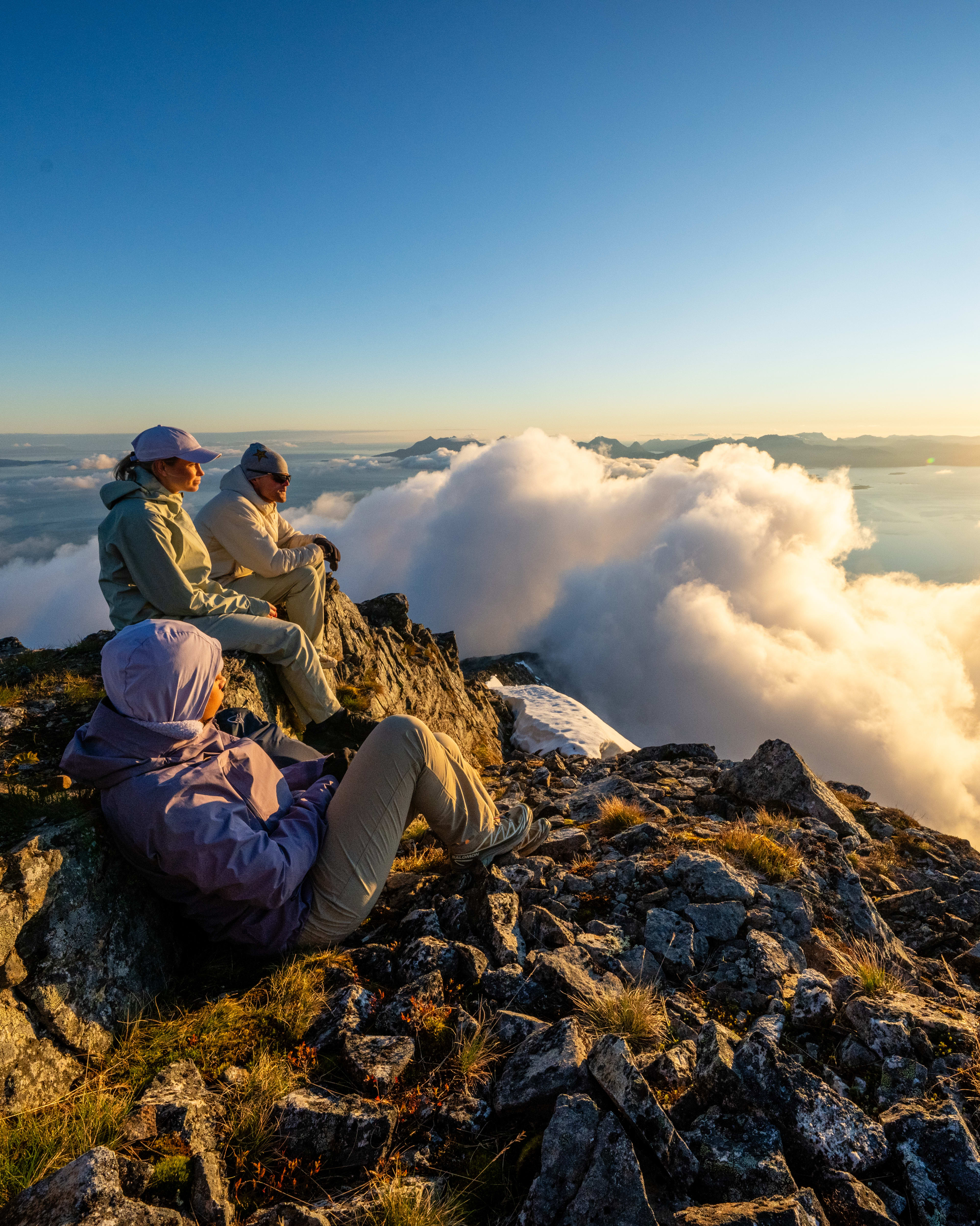 Three hikers in Norrøna clothing watch a sunset over clouds from a rocky peak.