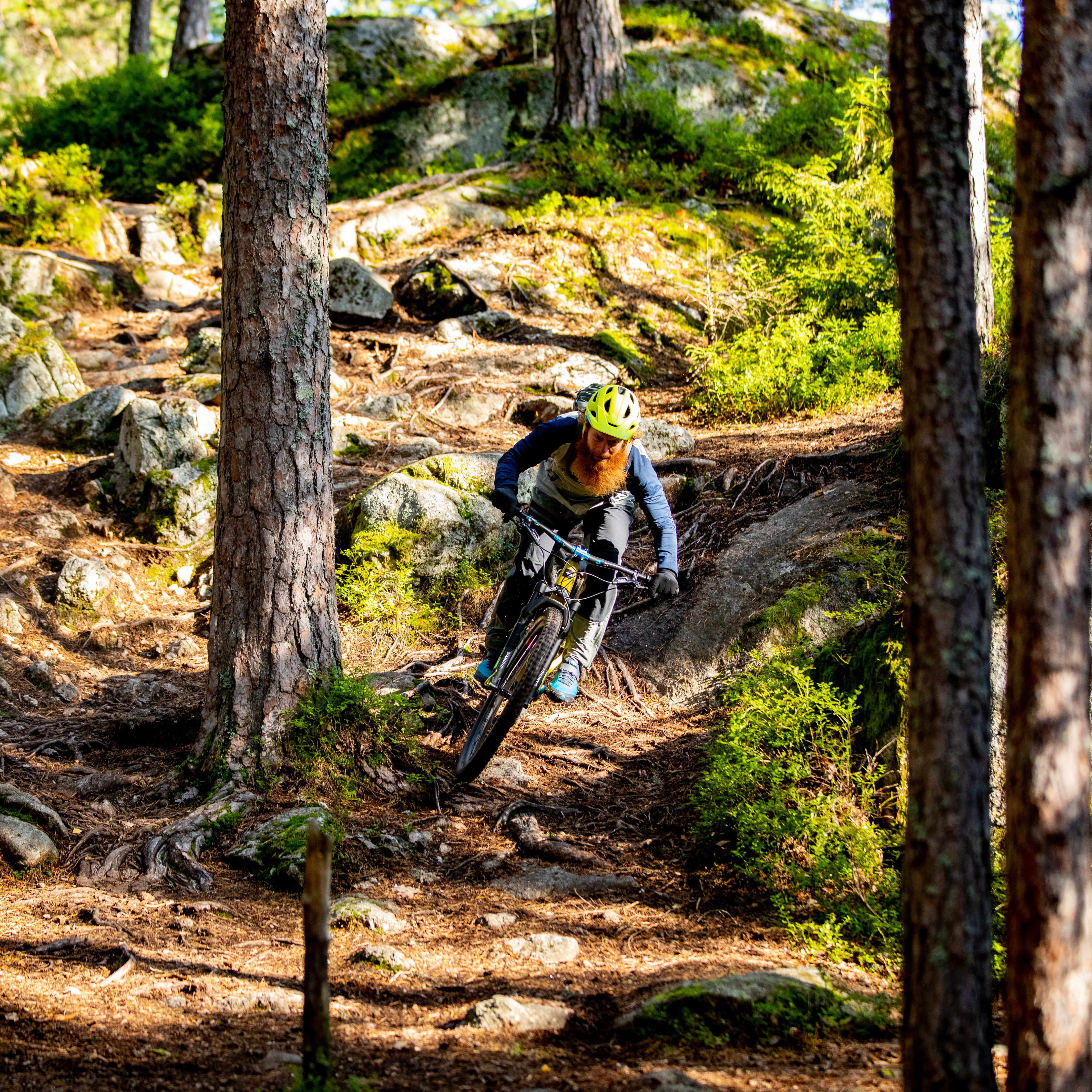 A man riding a mountain bike down a rocky path in a forest. He is wearing a helmet and Norrøna clothing. 
