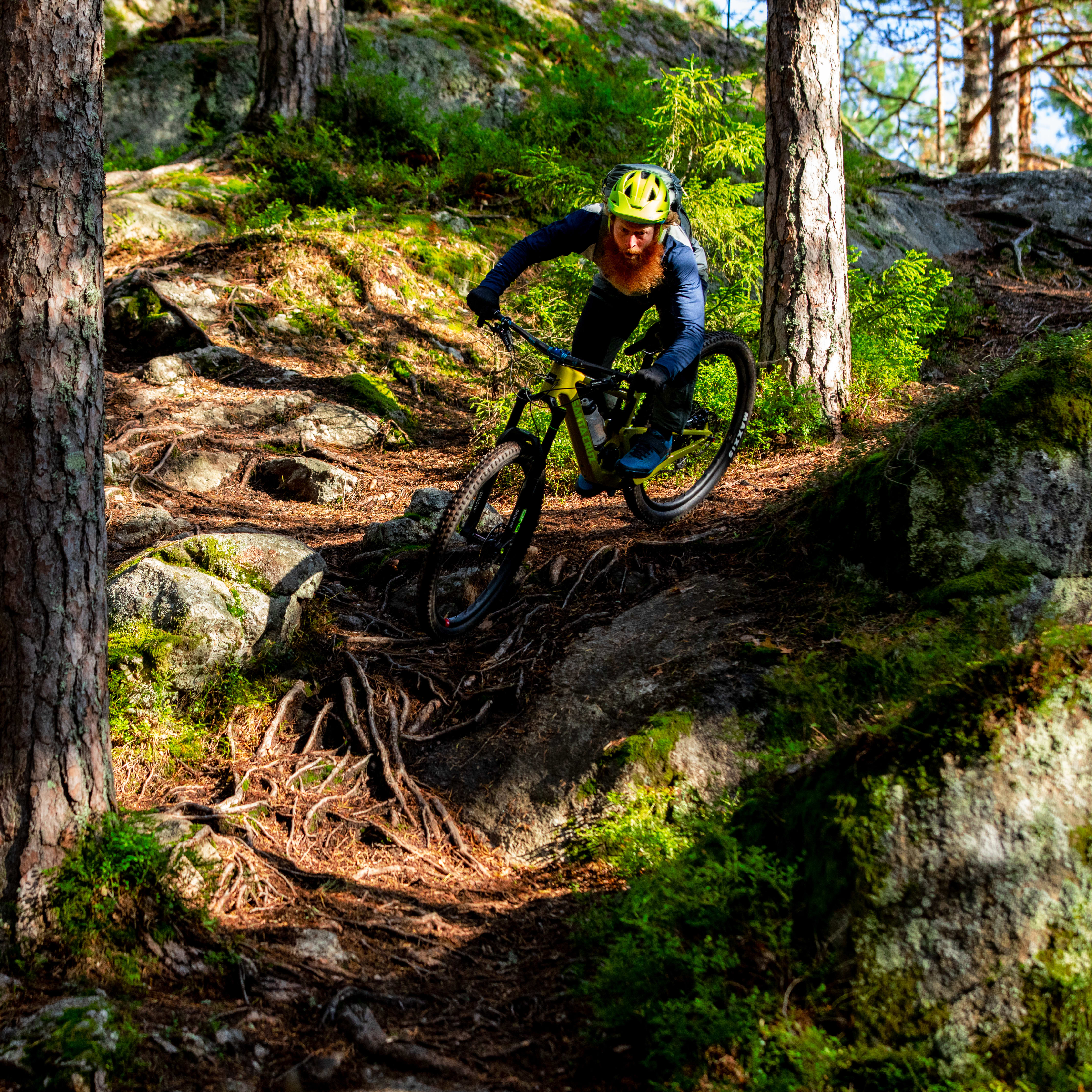 A person wearing a Norrøna cycling outfit rides a mountain bike on a rocky trail through a forest. 
