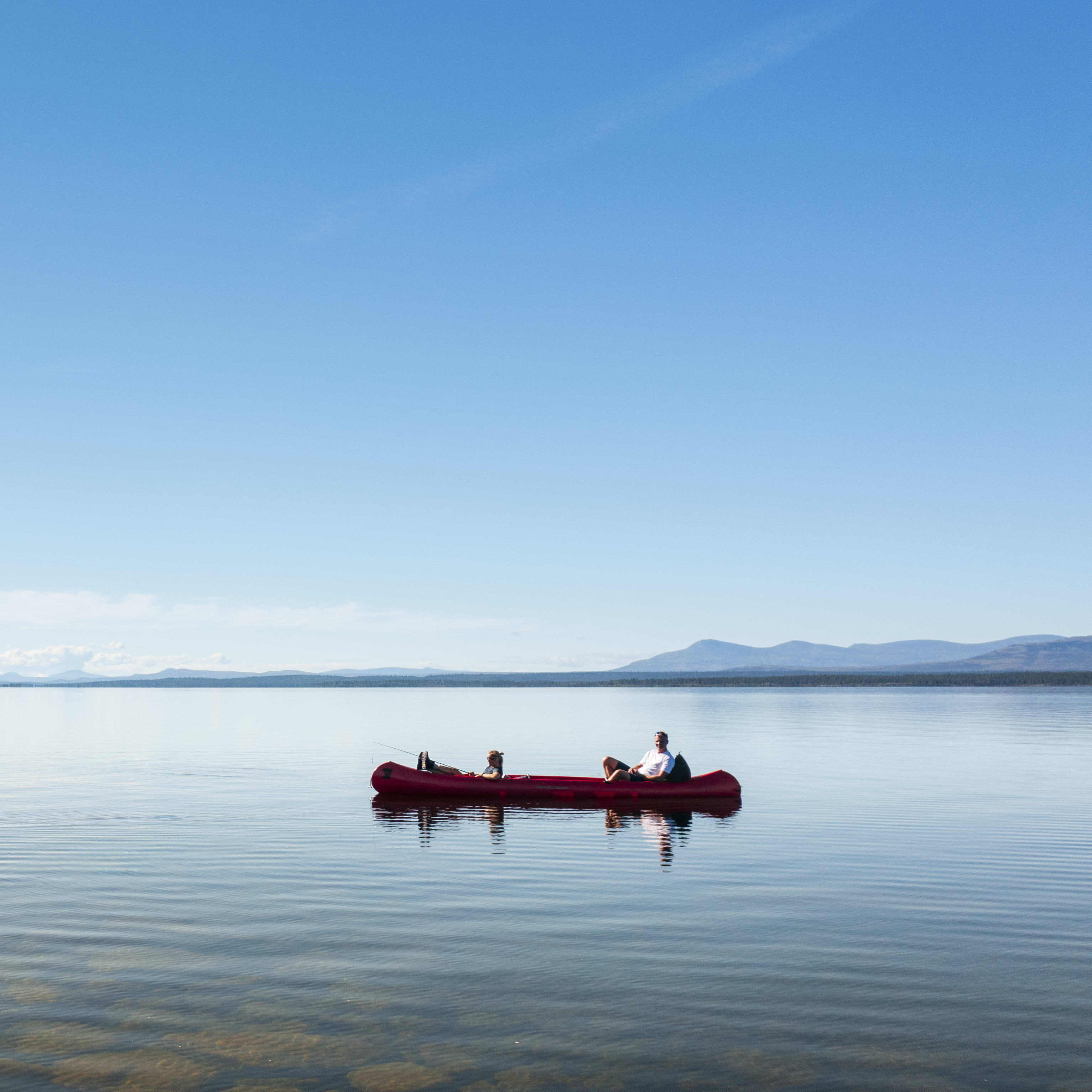 A person paddling a red canoe on a calm lake with clear water.  A small island with trees is visible in the background. 

