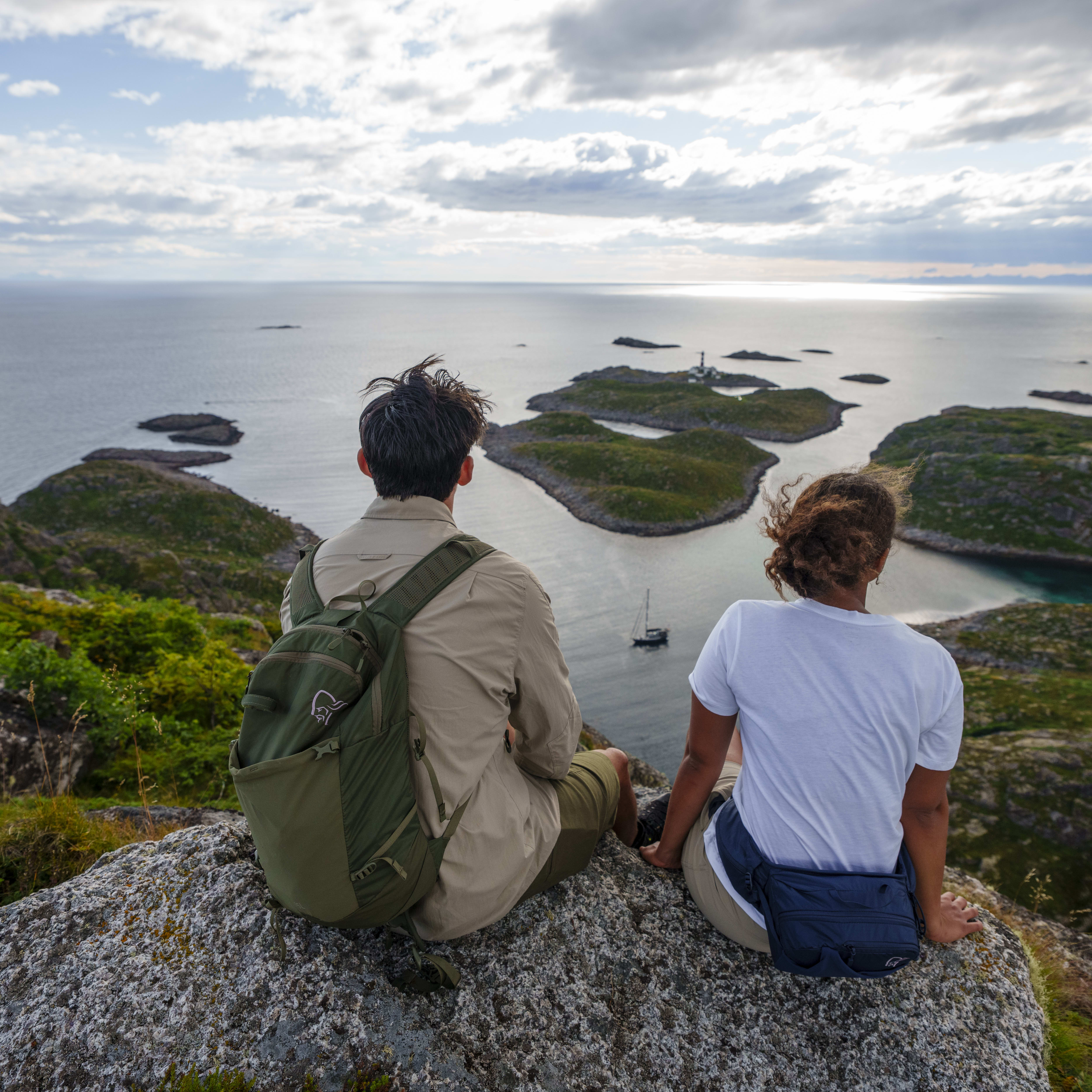 Two people sit on a rock, backs to the camera, overlooking a scenic island view. One wears a Norrøna backpack.

