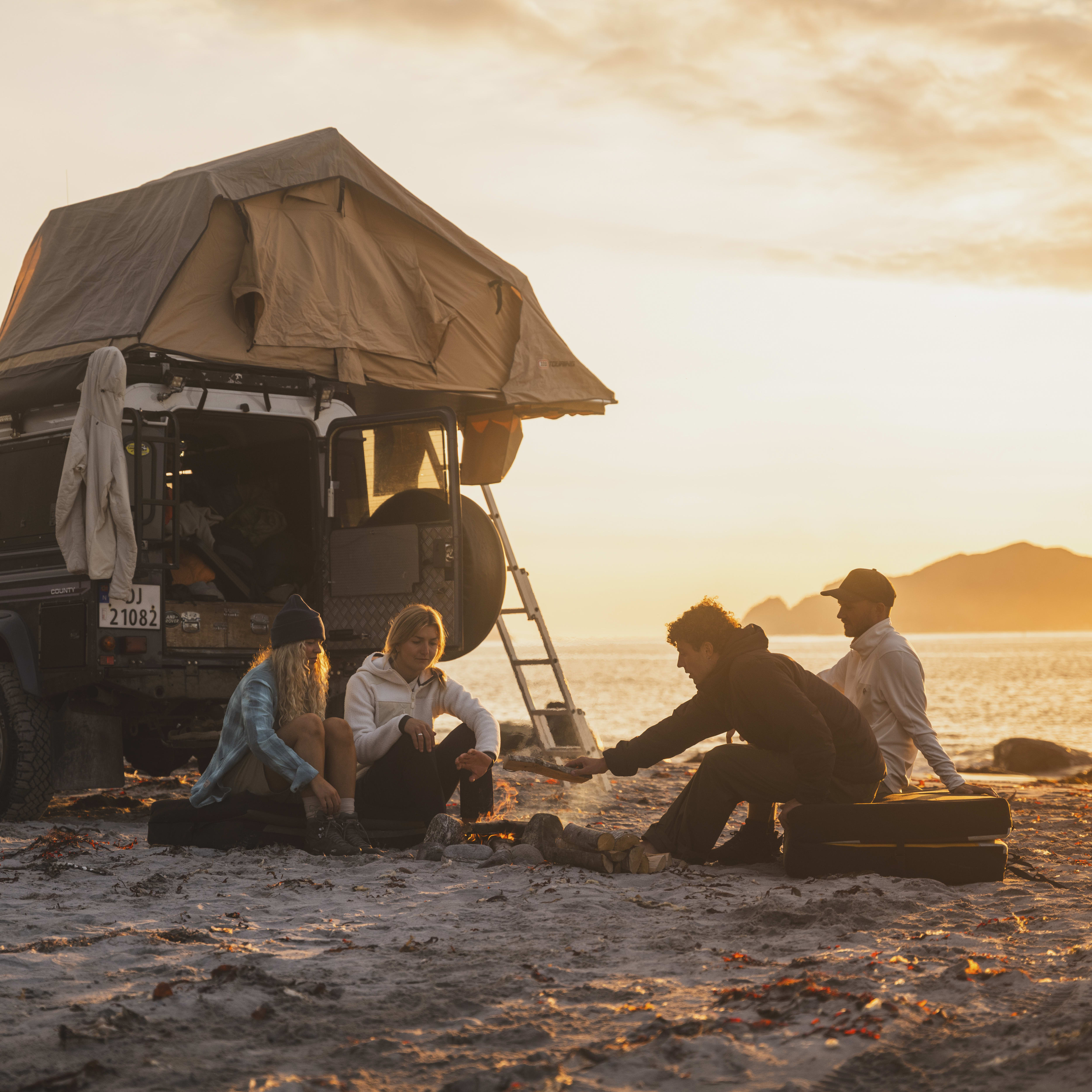 Four people sit around a campfire on a beach at sunset next to a black SUV with a roof tent. One woman wears a blue Norrøna flannel shirt.

