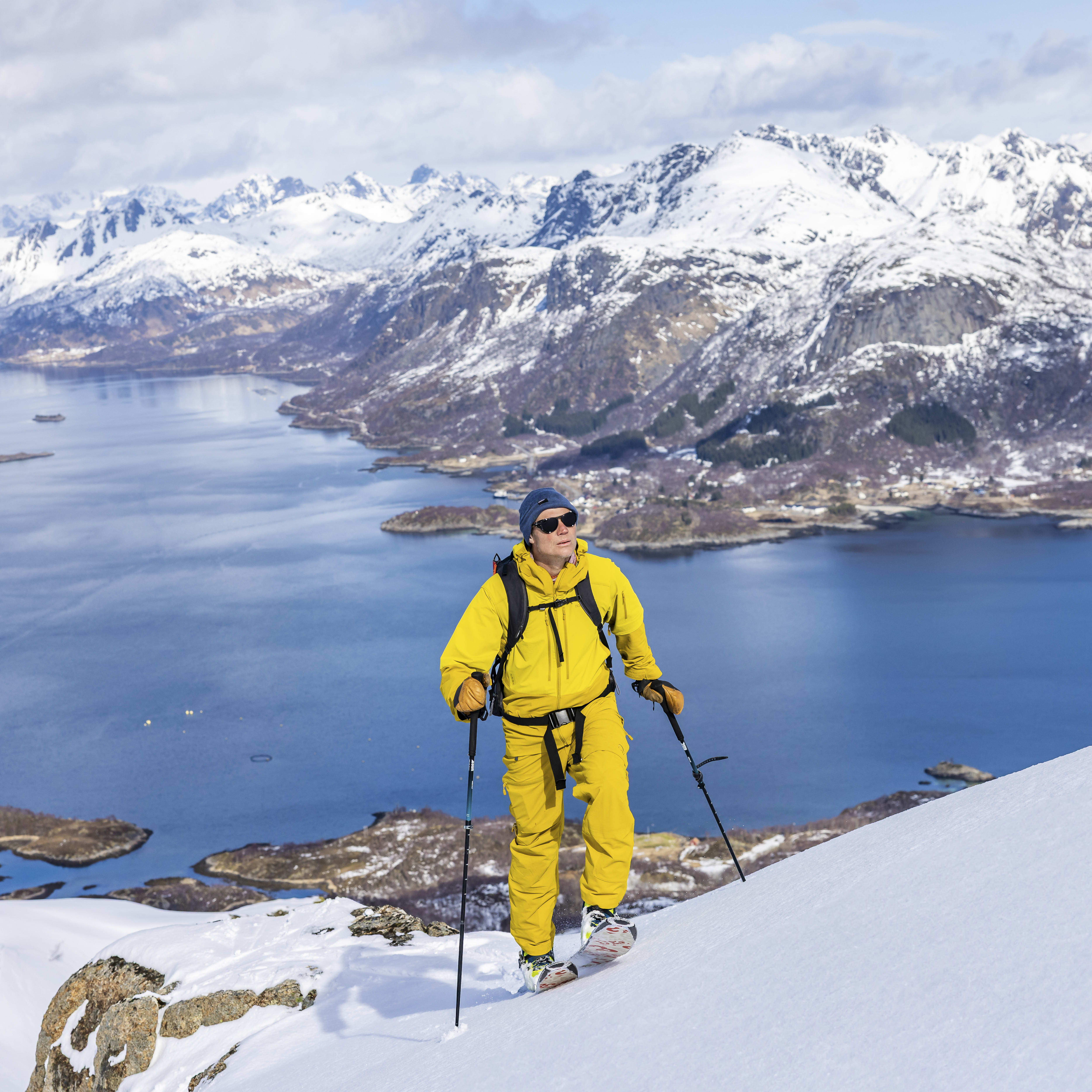 A person wearing a yellow Norrøna ski suit and sunglasses skiis down a snow covered mountainside overlooking a fjord.