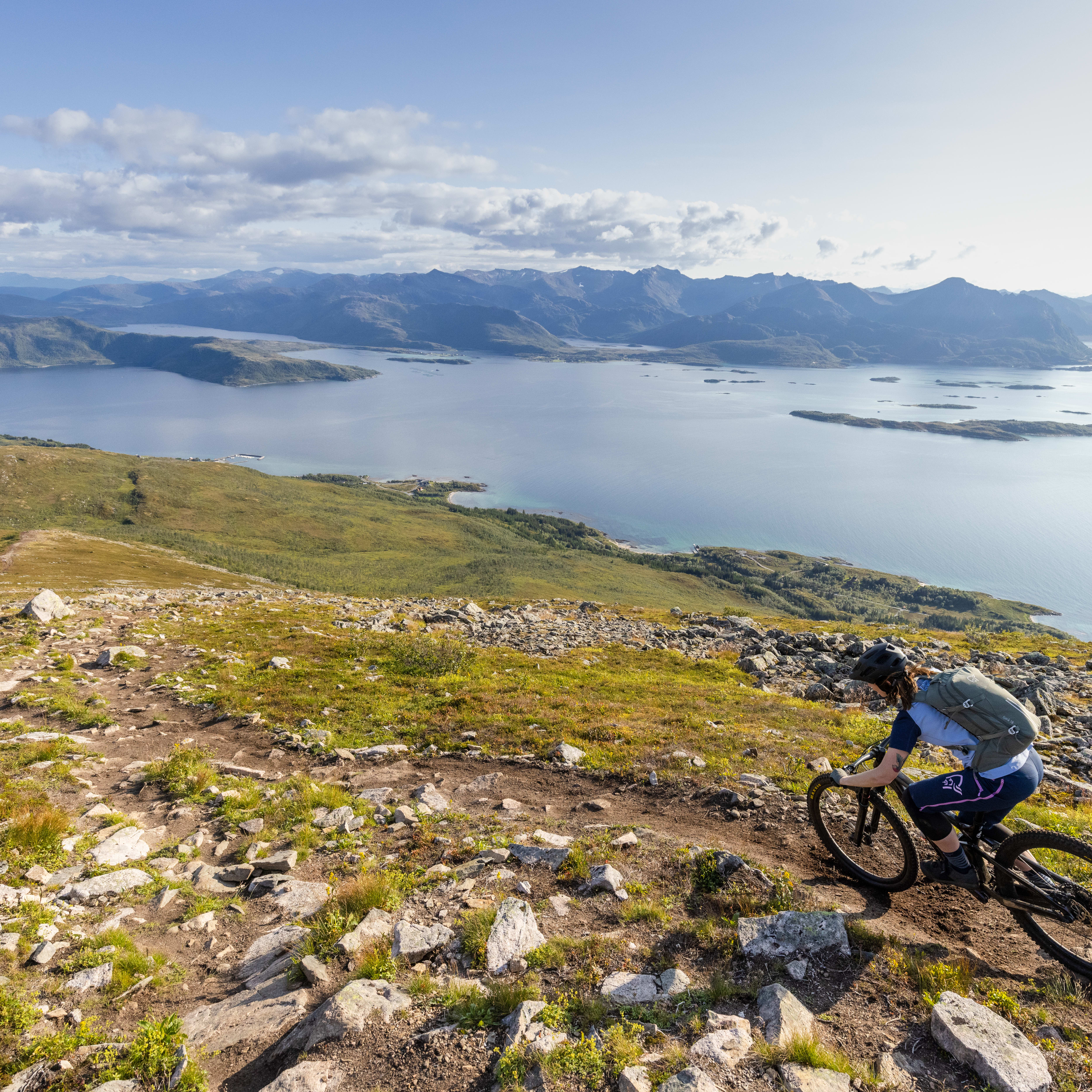 A person wearing a Norrøna shirt and a helmet rides a mountain bike down a rocky path with a scenic view of a body of water and mountains in the background. 
