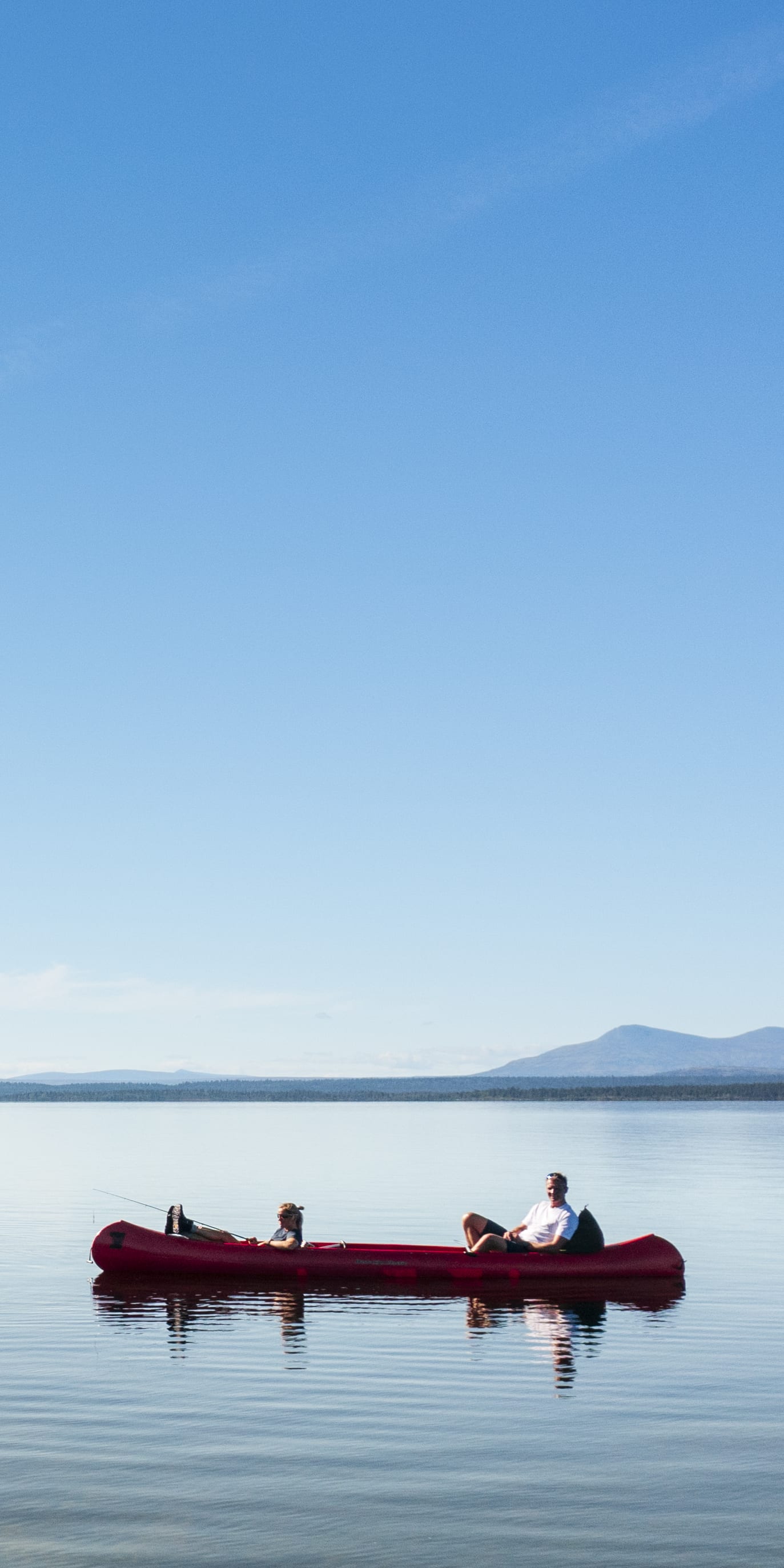 A person paddling a red canoe on a calm lake with clear water. A small island with trees is visible in the background.