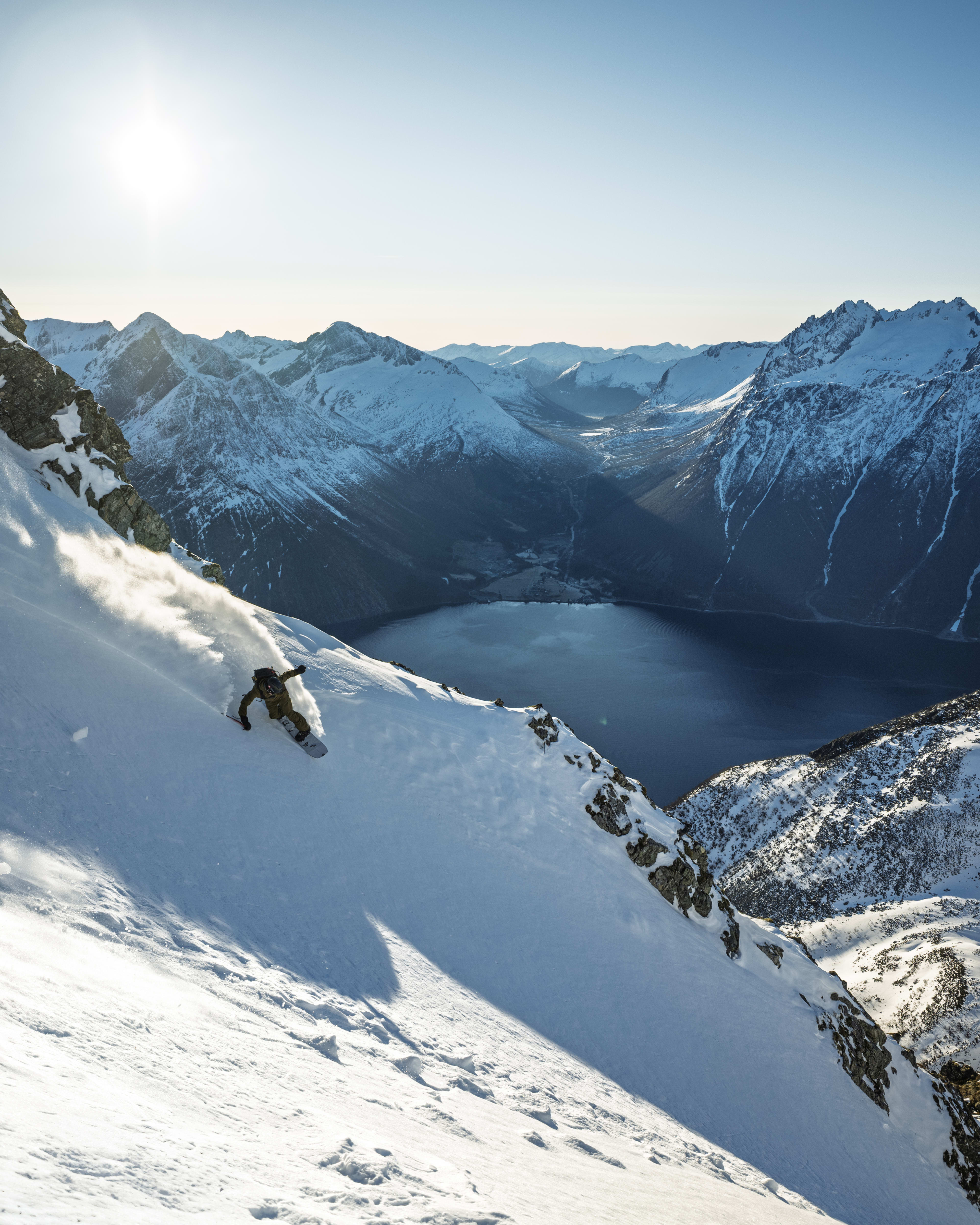 A snowboarder in Norrøna outerwear carves down a snowy mountain slope on a sunny day, with stunning mountain views in the background.