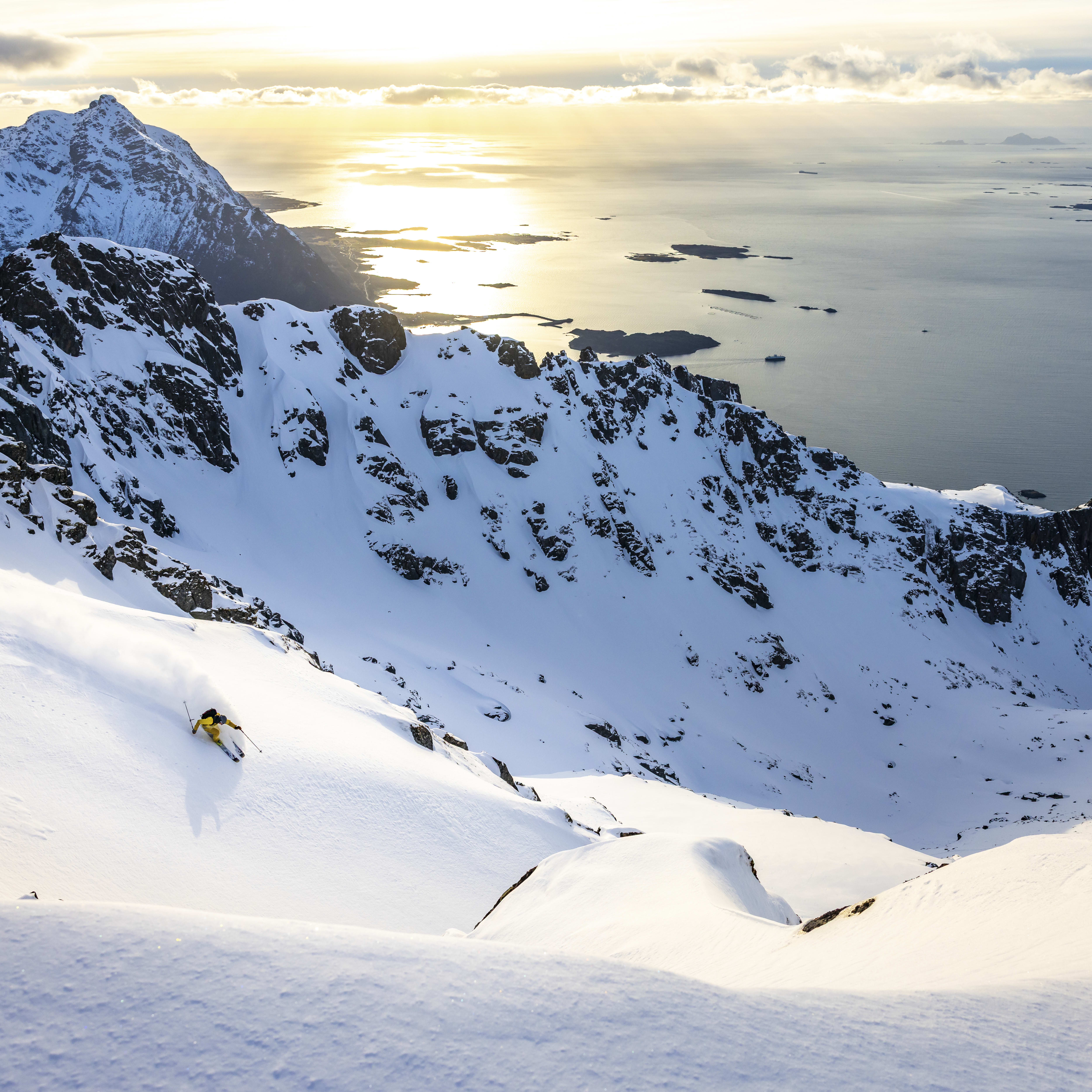 A skier in yellow clothing descends a snow-covered mountainside. The mountains overlook a vast, blue ocean in the distance. 
