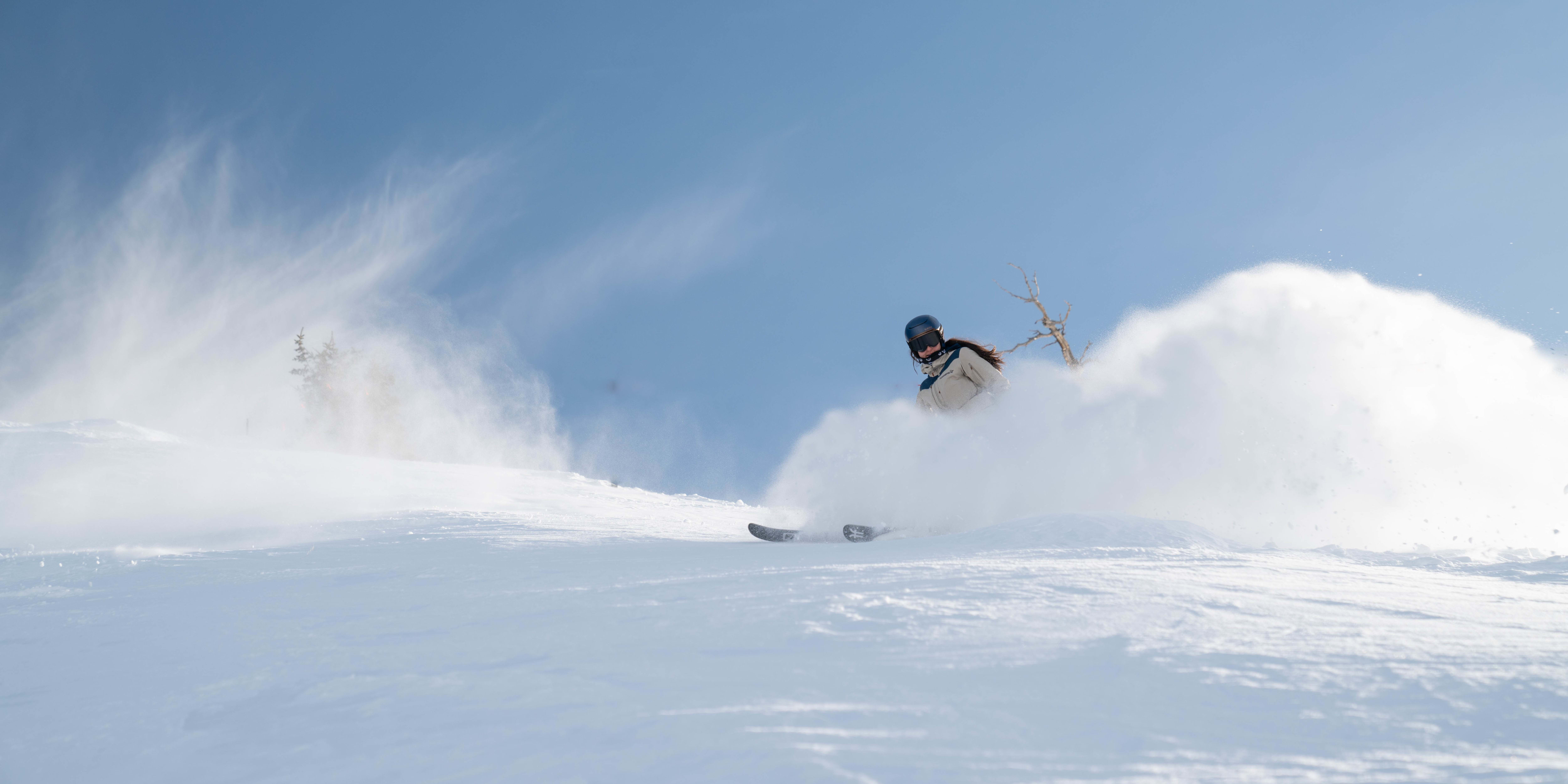 A skier in a Norrøna jacket carves through deep powder snow, kicking up a white spray against a clear blue sky.