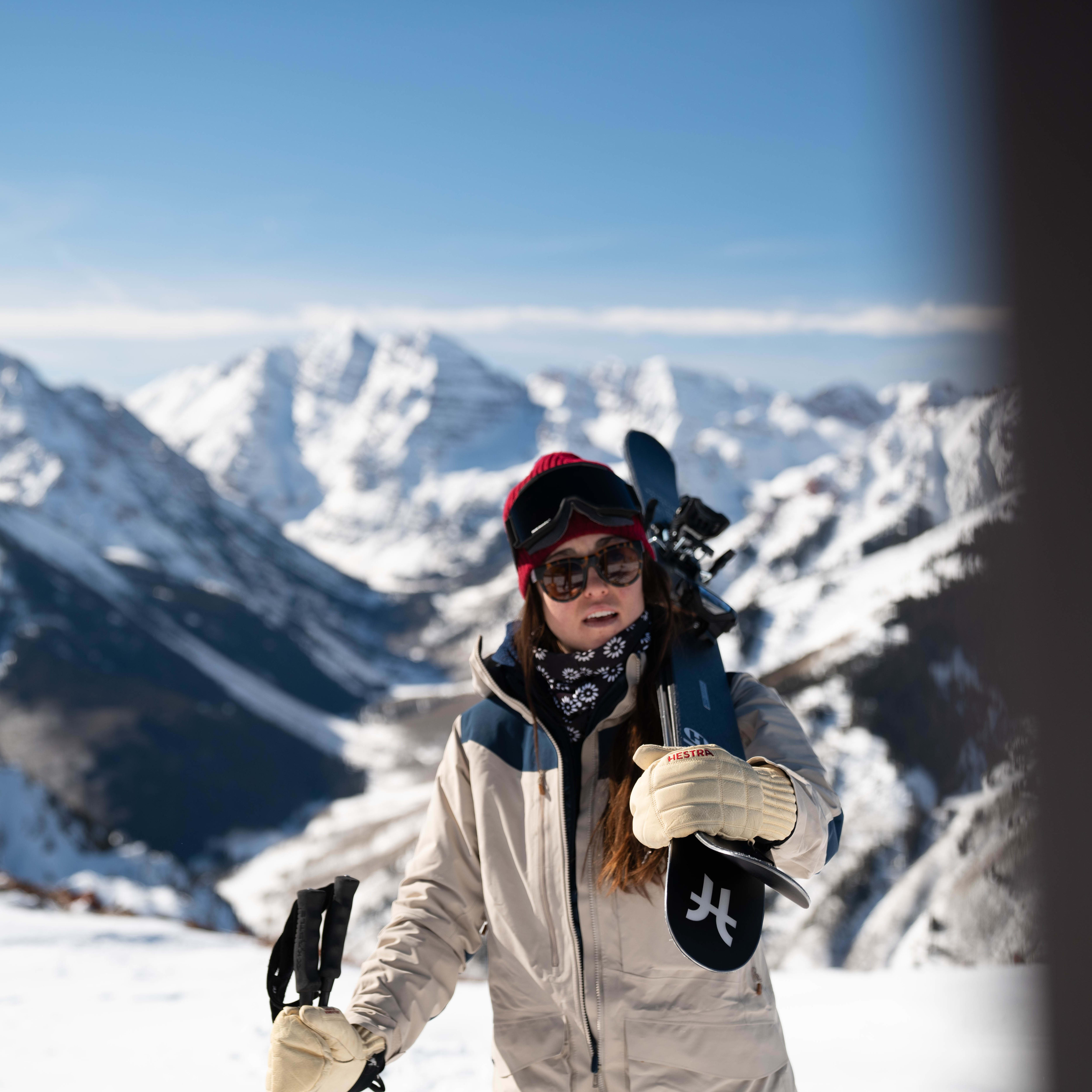 A skier in a Norrøna jacket and red beanie, carries skis with mountain backdrop under a clear blue sky.