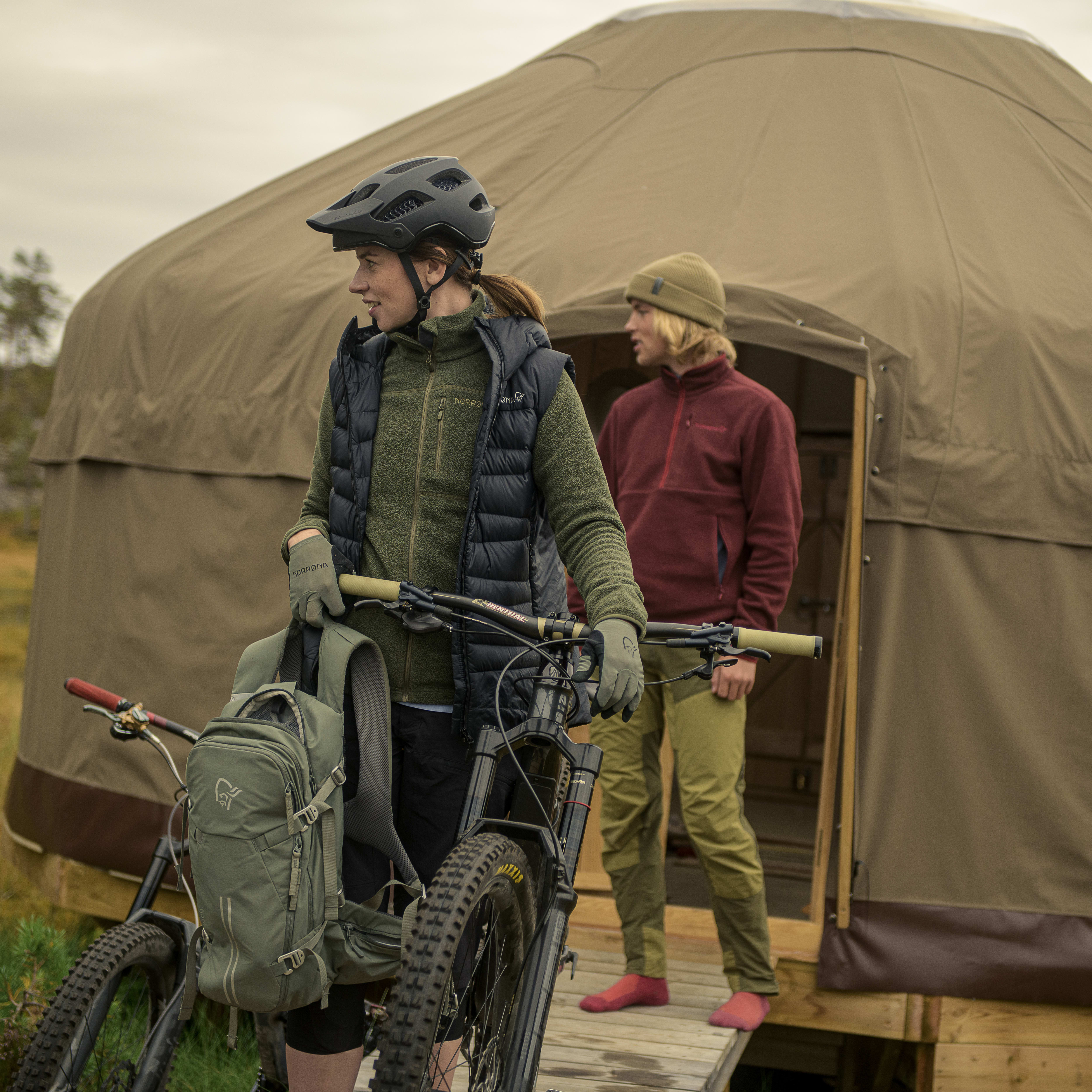 A woman wearing a Norrøna vest and a helmet stands next to a mountain bike.  The bike is in front of a large tent.