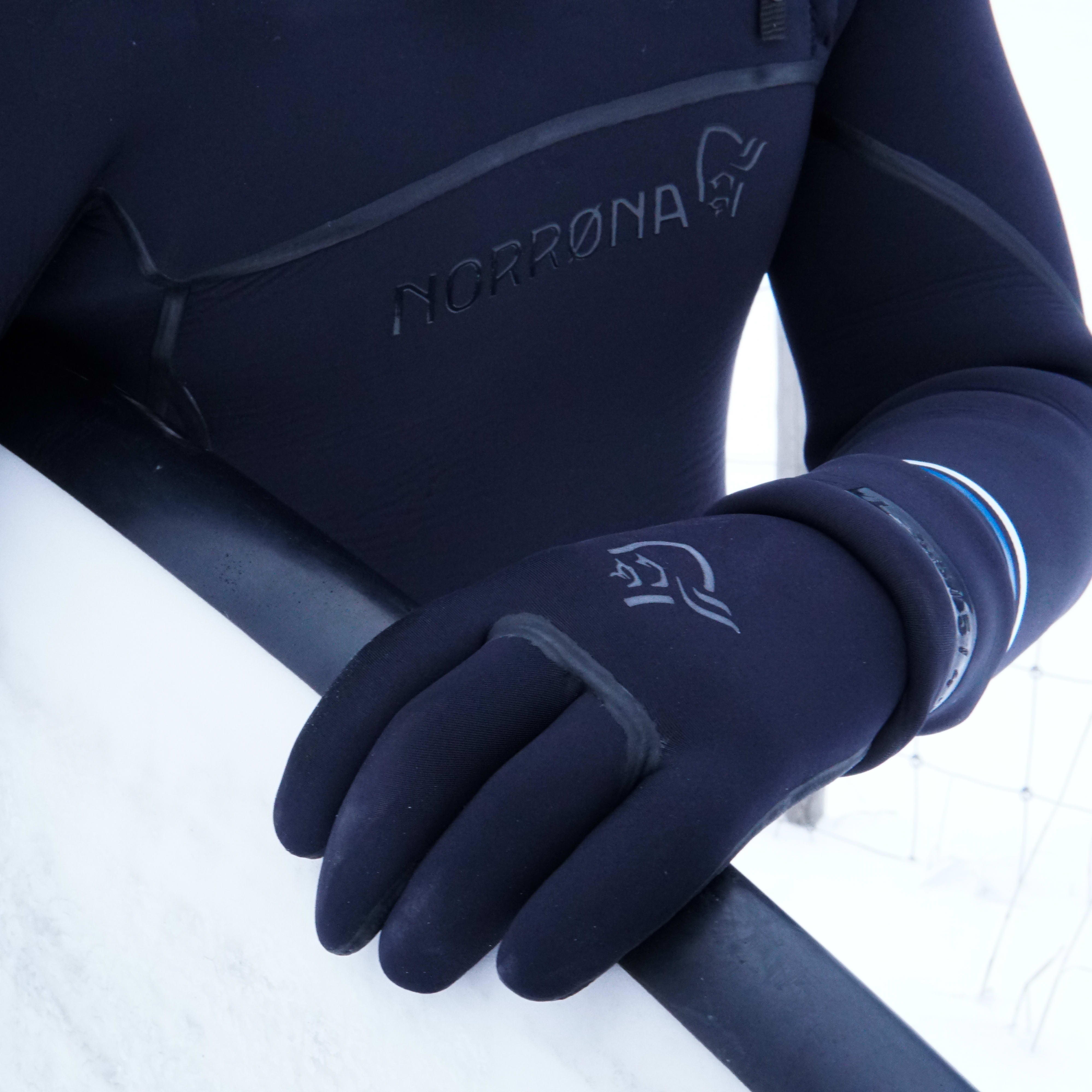 Person wearing a black Norrøna wetsuit and gloves holding a white surfboard in snowy conditions. 
