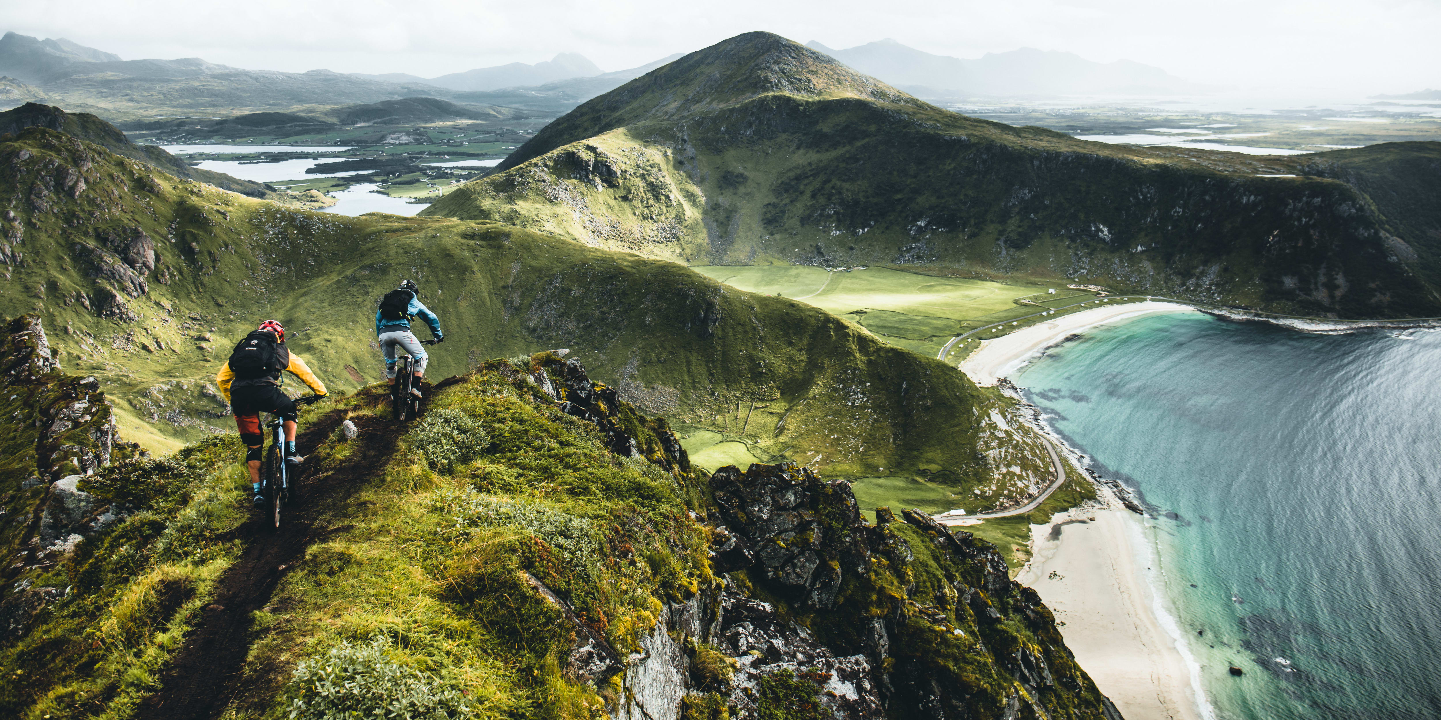 Two people mountain biking down a steep, grassy slope. The trail leads to a beach and a vast ocean view. 
