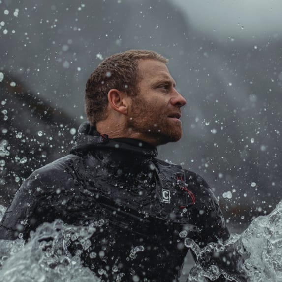 A man in a black Norrøna wetsuit stands in splashing water, looking to the side. A blurred mountainous background is visible.

