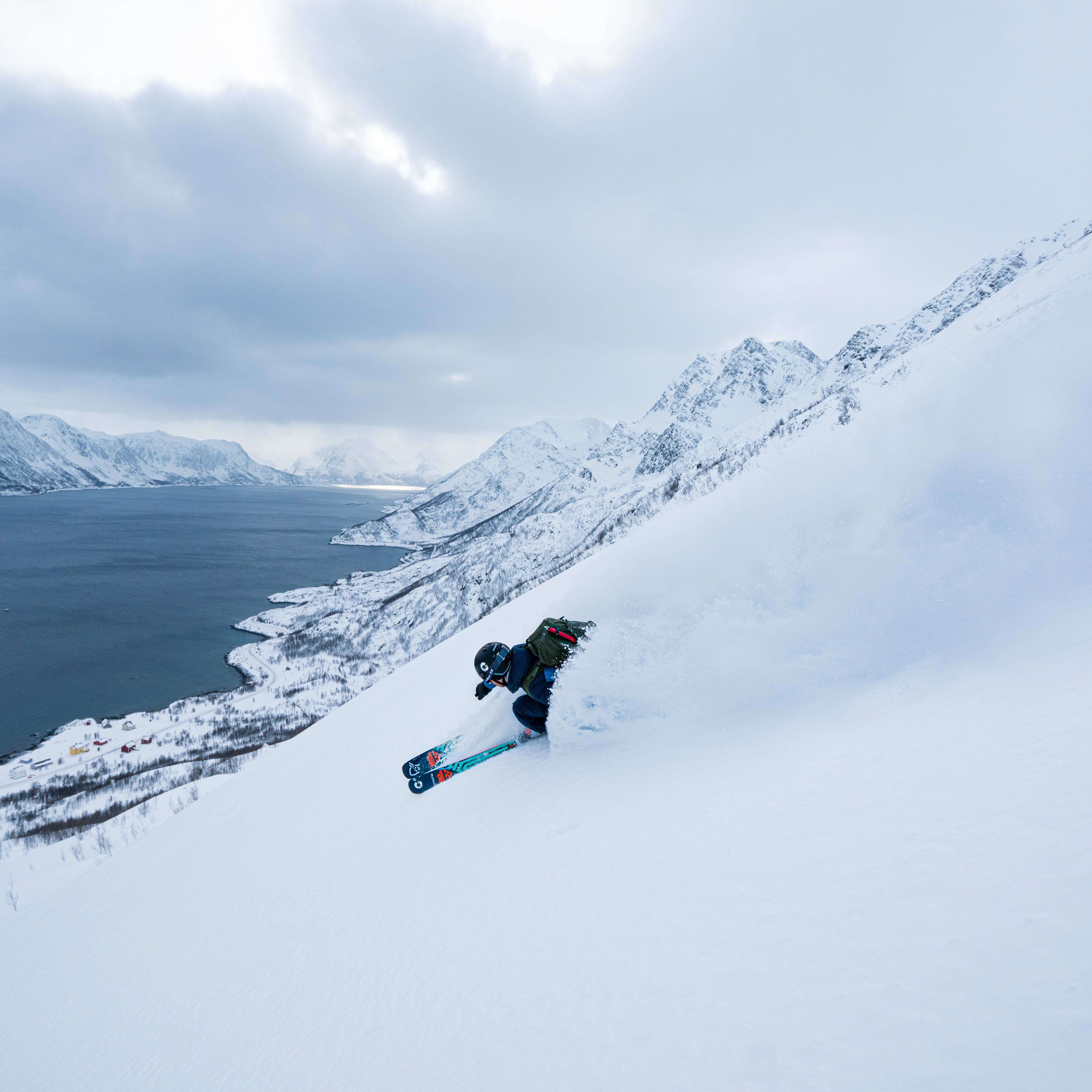 A skier wearing Norrøna gear descends a snowy mountain slope, with a fjord and distant mountains in the background.