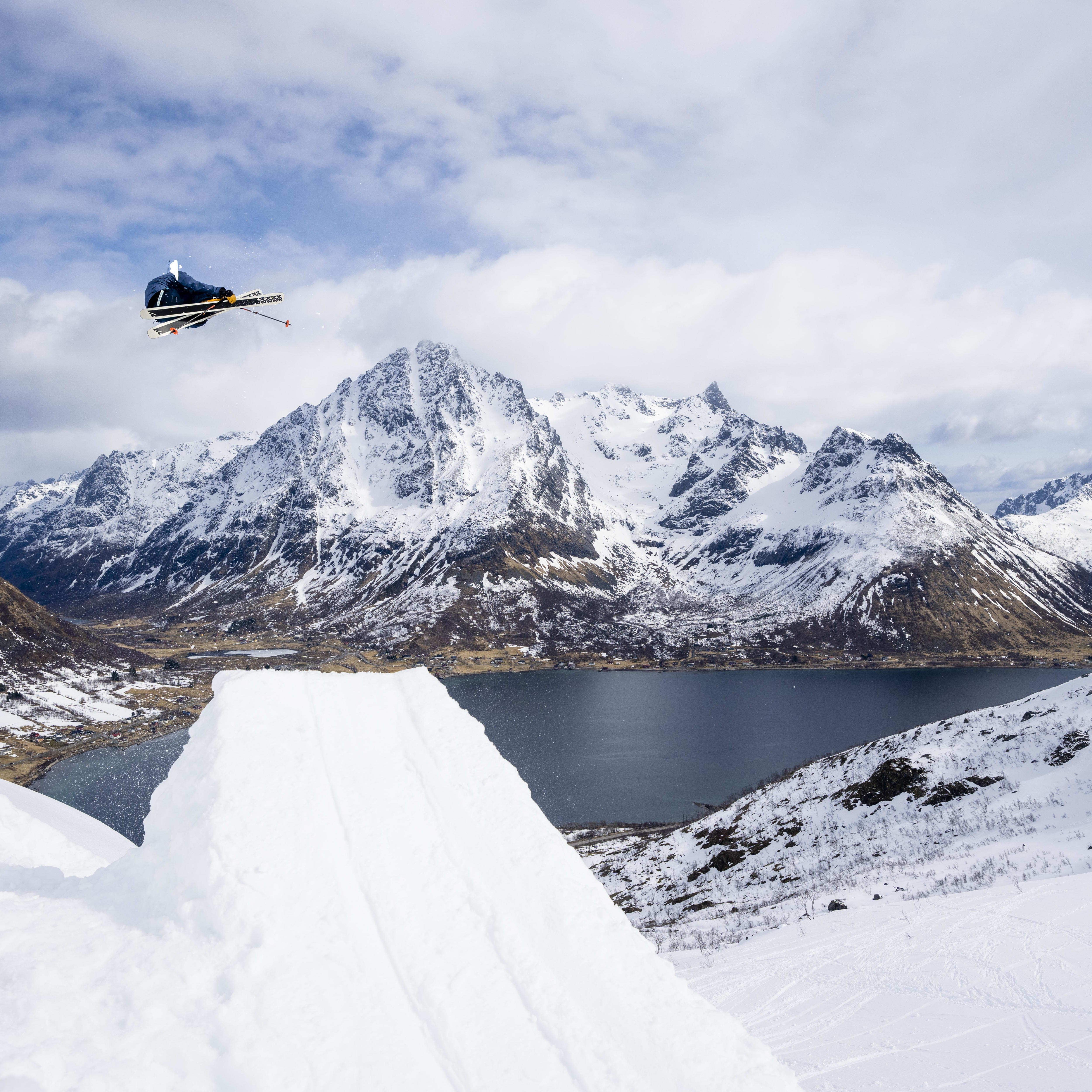 A skier wearing Norrøna clothing performs a trick off a snow jump. The skier is silhouetted against a blue sky with white clouds. In the background, a snowy mountain range and a fjord can be seen.
